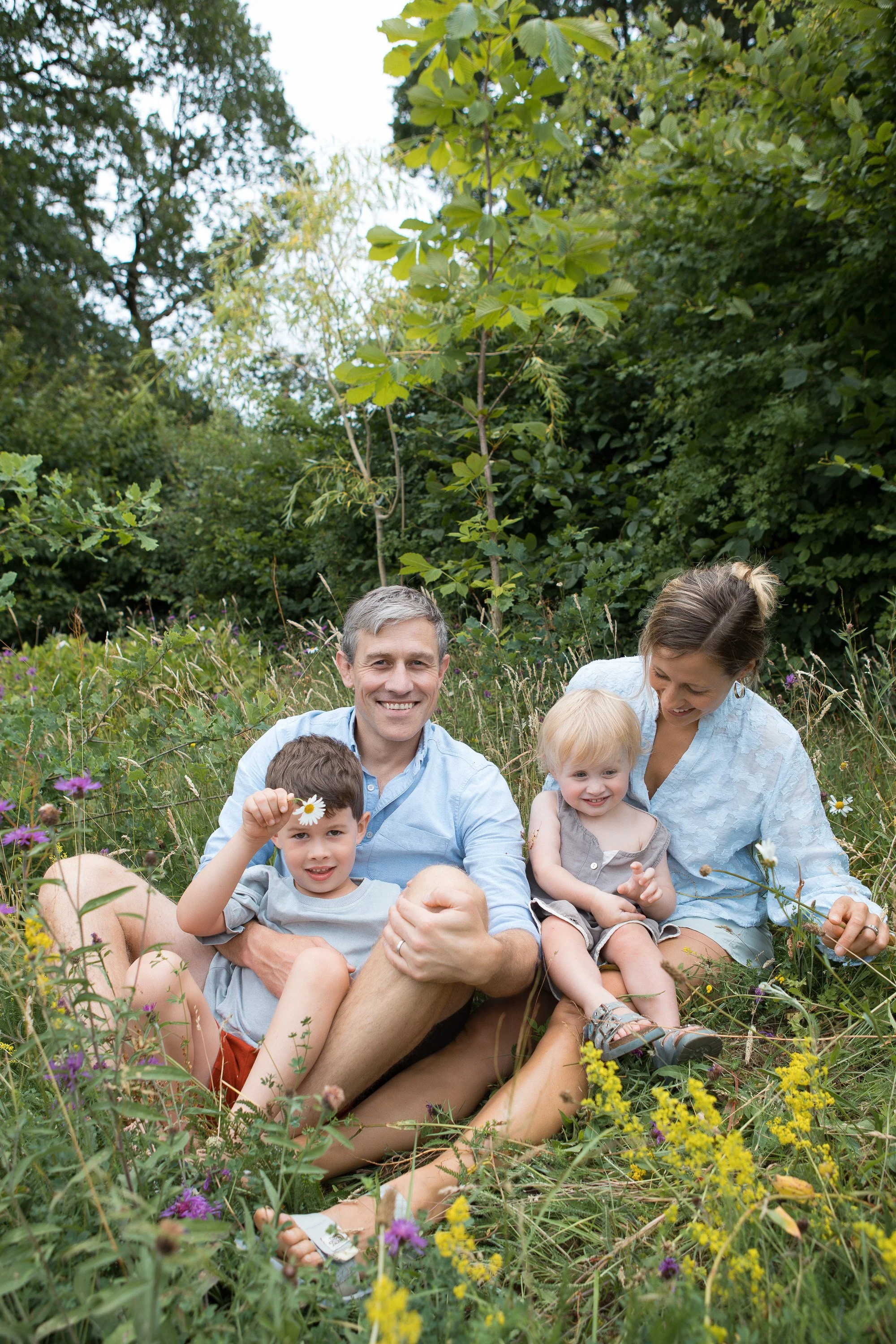 A family of five sitting in a lush, green outdoor meadow surrounded by wildflowers with trees and bushes in the background. The father is smiling and holding a child with a daisy, while the mother and other children sit close together.