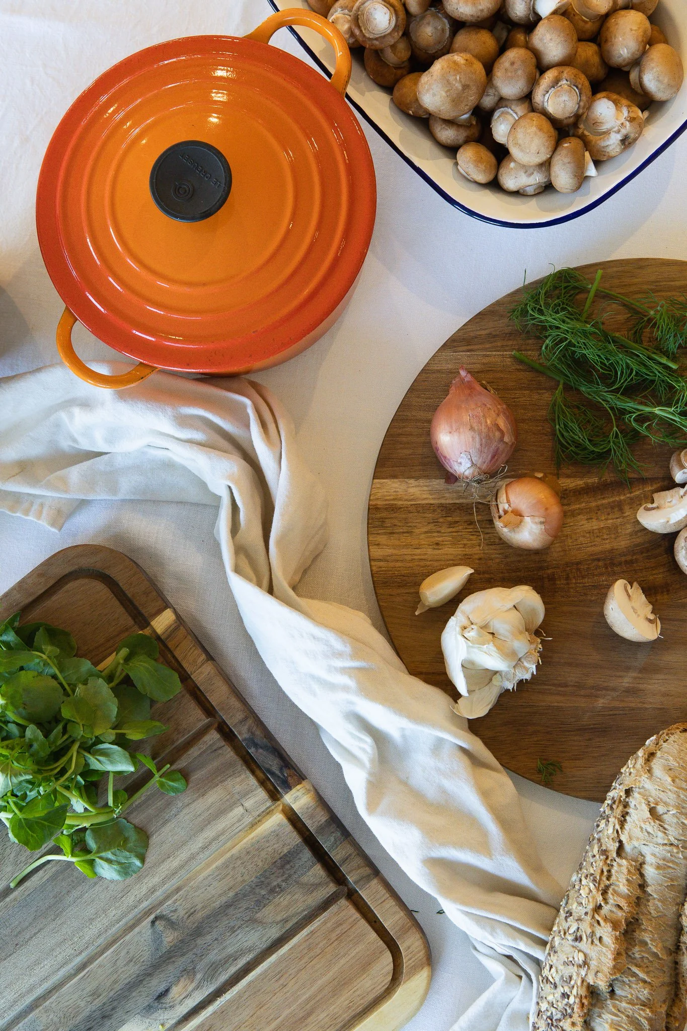 Top-down view of a kitchen countertop with a round wooden cutting board holding fresh herbs, two onions, and garlic cloves. To the right, a white dish filled with mushrooms. To the left, a wooden tray with green leafy herbs. A white cloth drapes acro