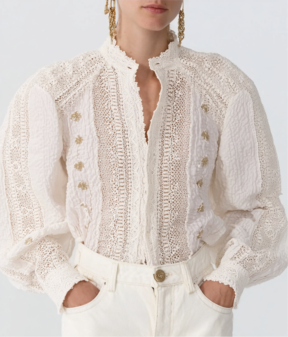 A woman wearing a white, intricately lace embroidered blouse with puffy sleeves and gold button details, paired with white pants and gold earrings.