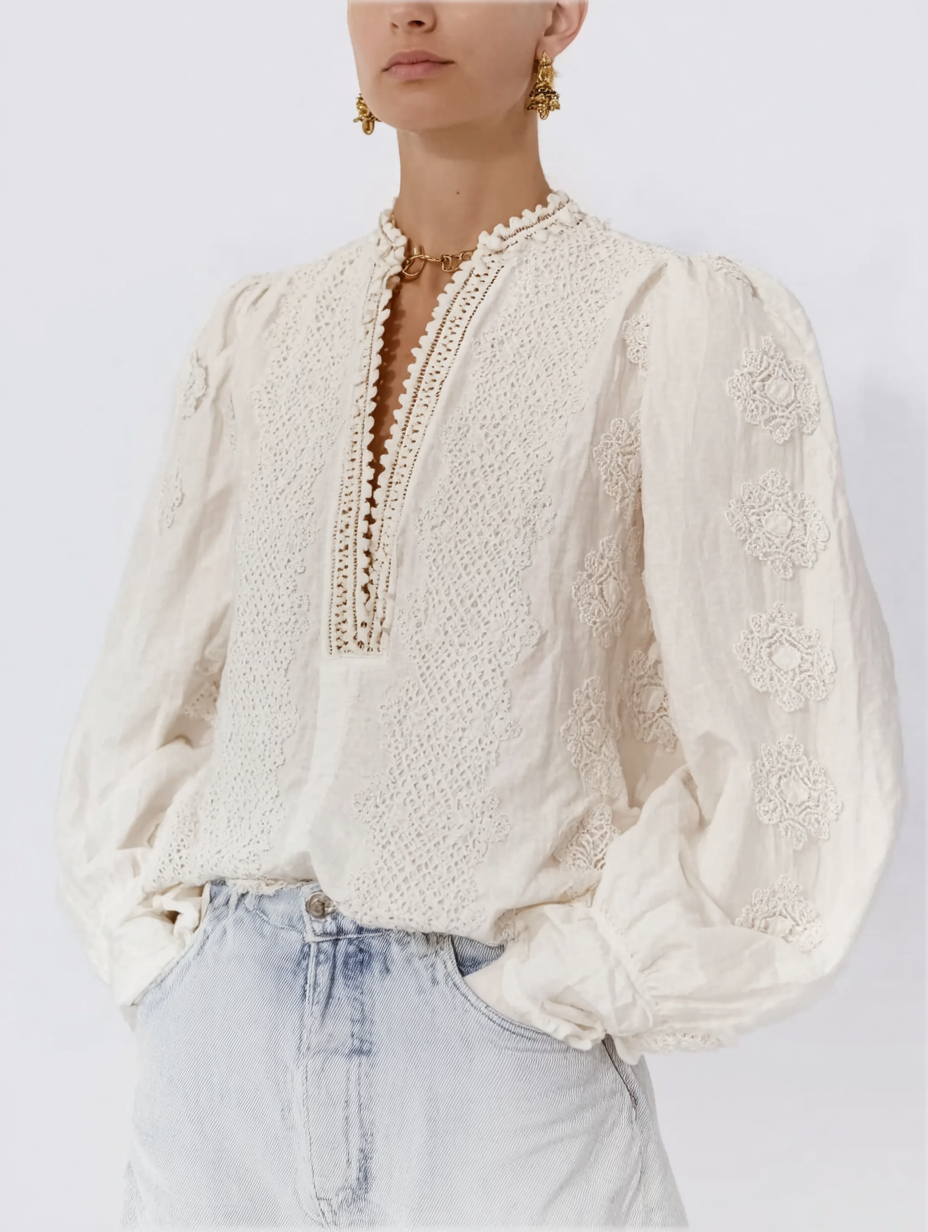 A woman wearing a cream-colored embroidered blouse with puff sleeves, light-washed jeans, gold earrings, and a gold chain necklace.