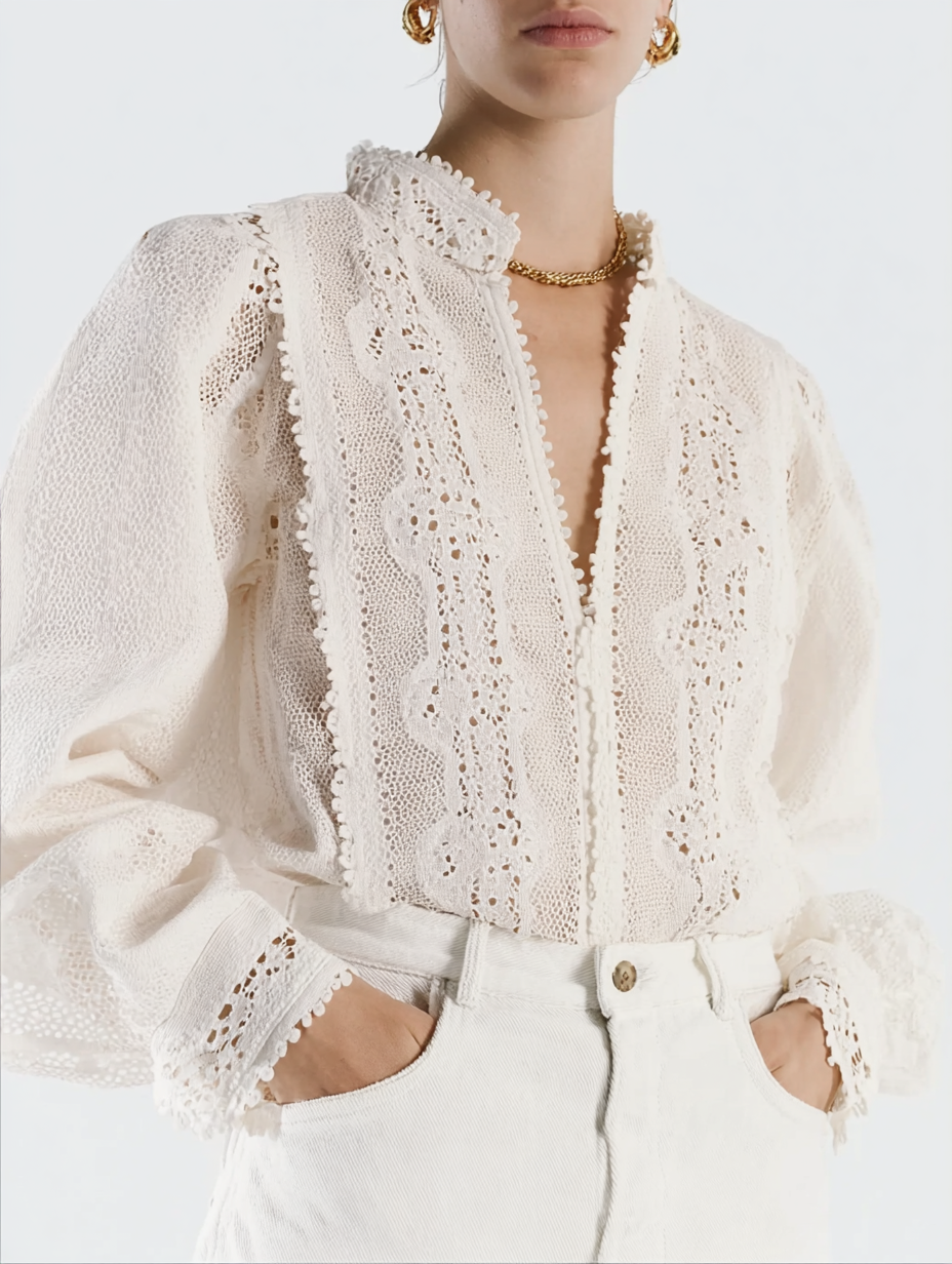 A woman wearing a cream-colored lace blouse with intricate embroidery, puffed sleeves, and a high neckline, paired with cream pants and accessorized with gold jewelry.