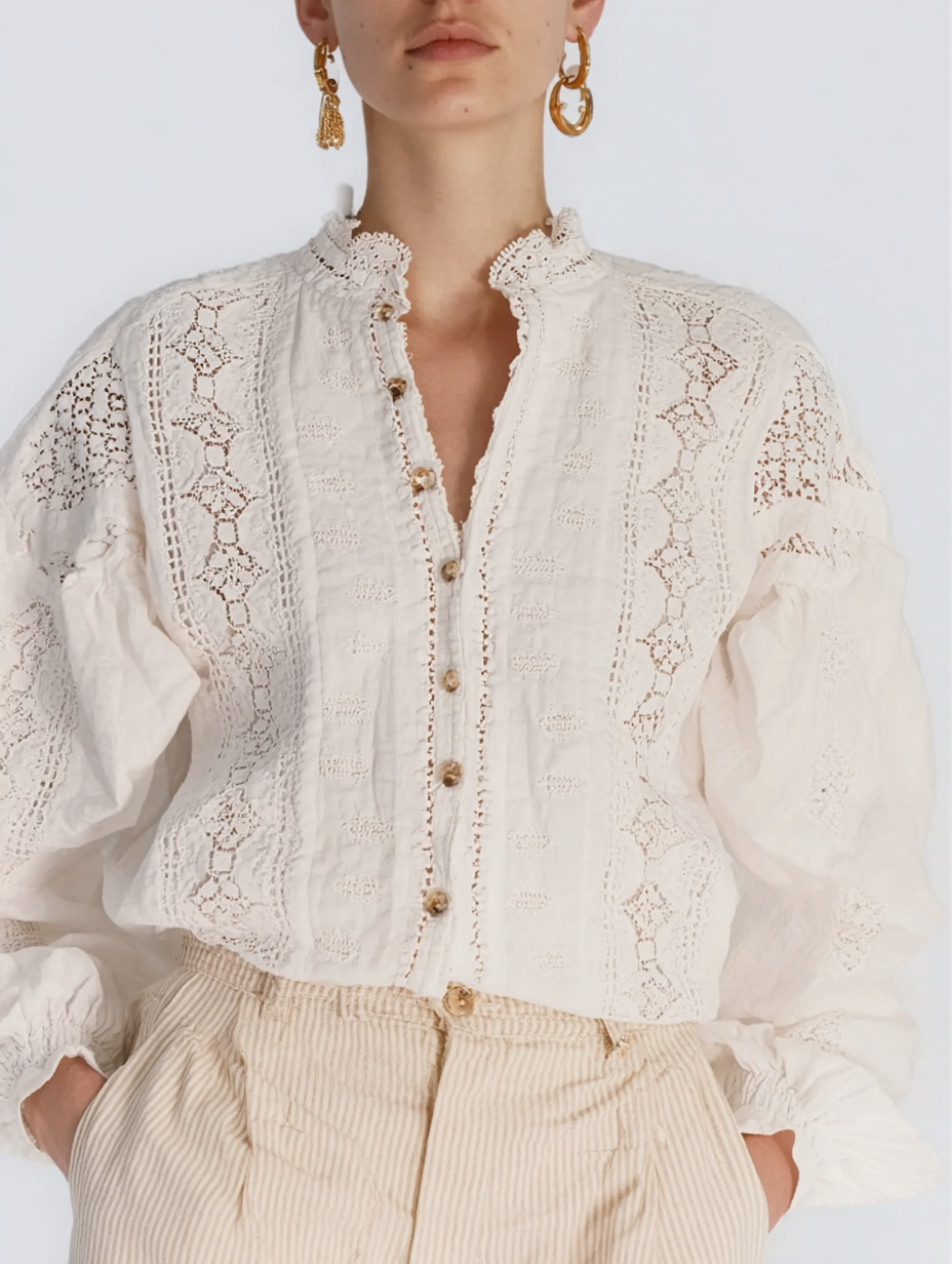 A woman wearing a cream-colored lace blouse with embroidered details and beige high-waisted pants, accessorized with large gold hoop earrings.