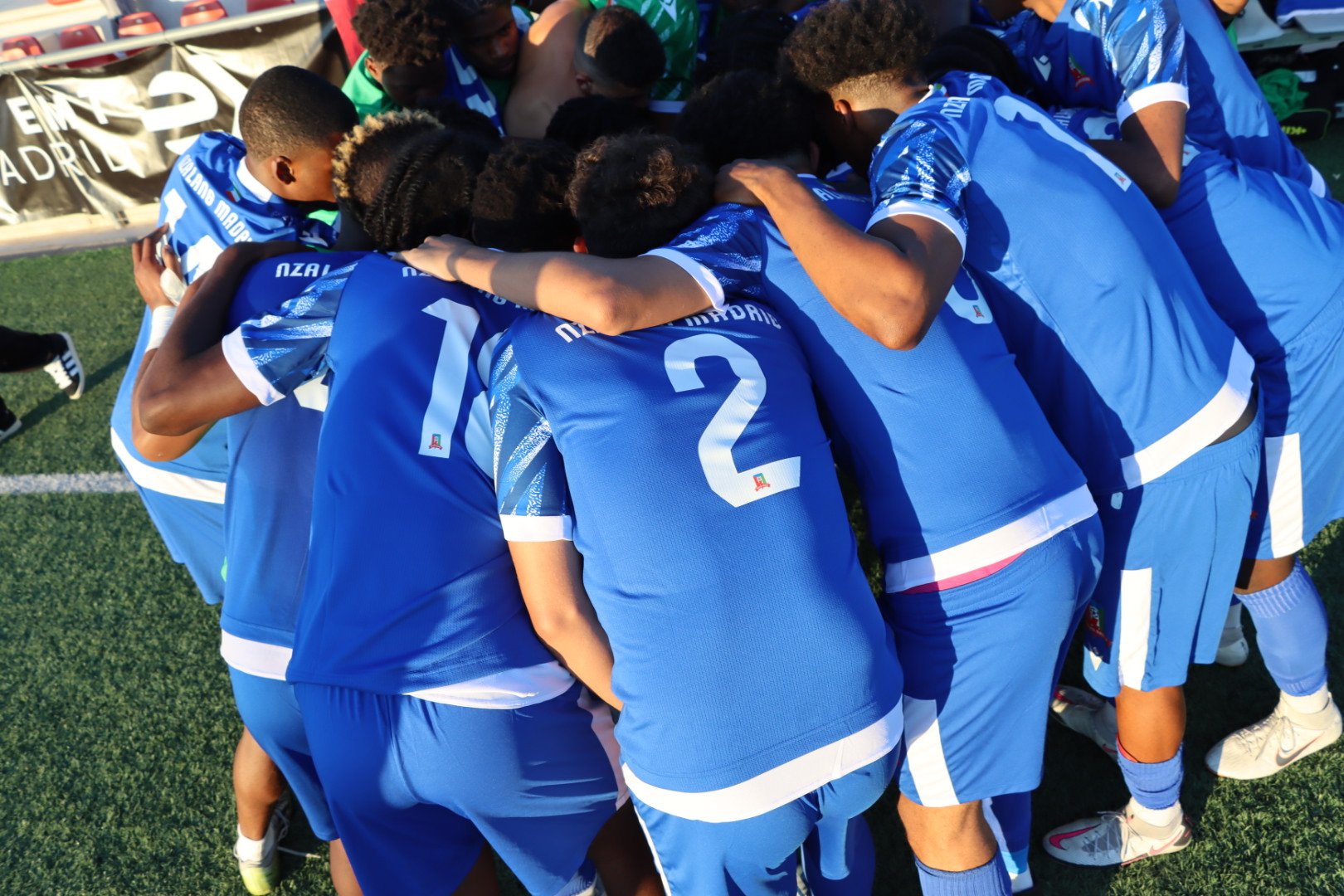 Grupo de futbolistas en uniforme azul formando un círculo y abrazándose en el campo.