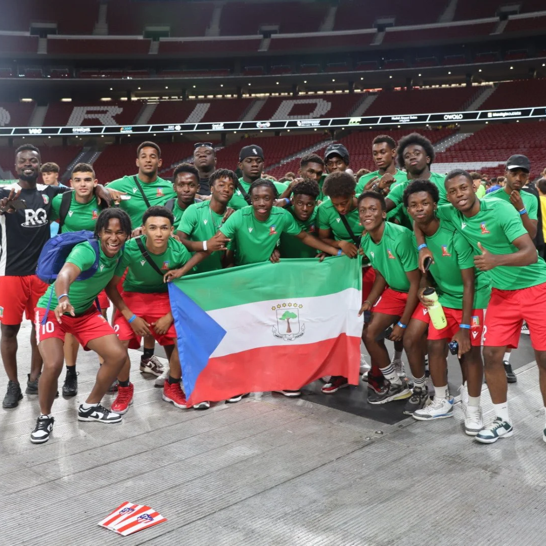 Grupo de jóvenes jugadores de fútbol en un estadio, sosteniendo una bandera de Django, con vestimenta deportiva verde y roja, celebrando después de un partido o evento deportivo.
