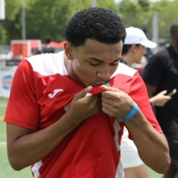 Un joven deportista con uniforme rojo y blanco besa su camiseta en un campo abierto.
