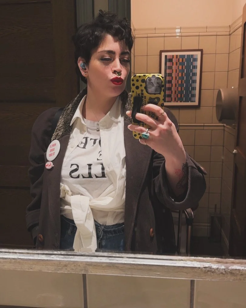 A woman with short dark hair taking a mirror selfie in a bathroom. She has bright red lipstick, multiple earrings, and a septum piercing. She wears a white shirt tied at the waist, a dark blazer, and jeans. She has a tattoo on her left forearm and several buttons pinned to her blazer.