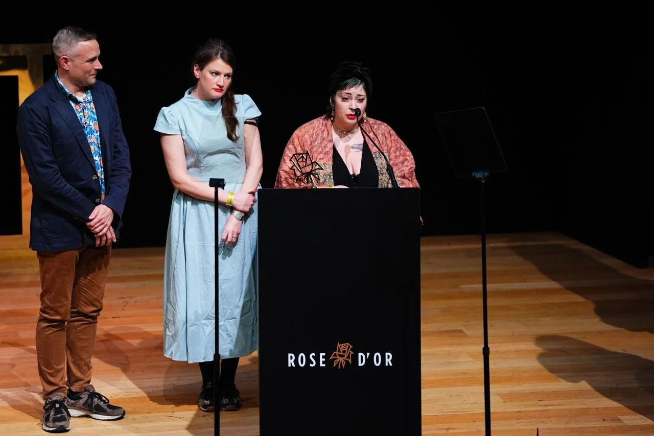 Three people standing on a wooden stage with a black backdrop, one woman speaking at a podium labeled 'ROSE D'OR,' and two people listening.