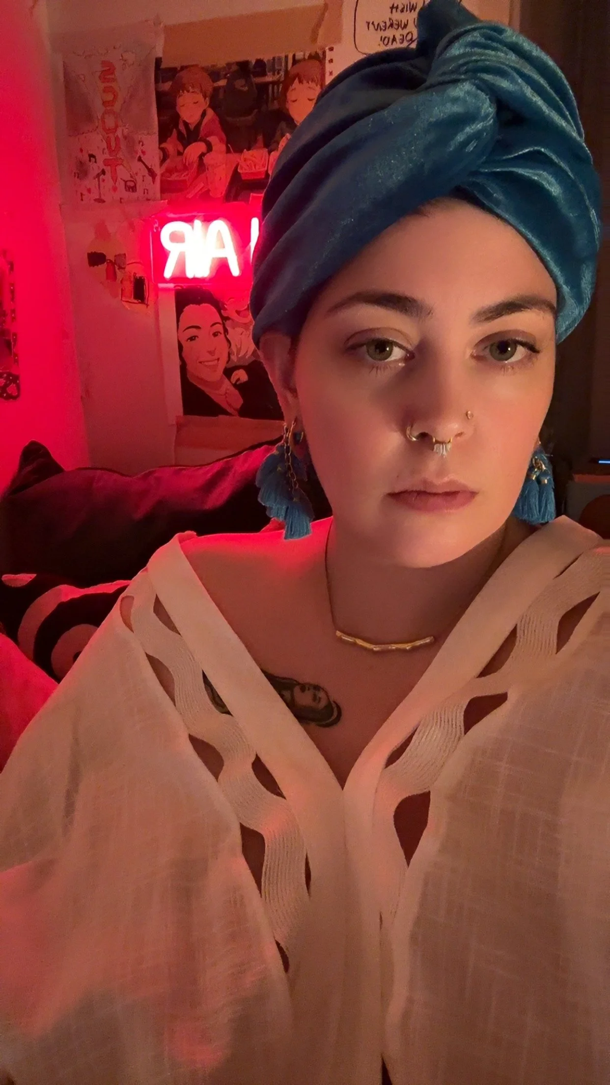 A young woman with short hair, wearing a blue head wrap, gold jewelry, and a beige top with cut-out details, sitting in a dimly lit room with pink and red lighting, decorated with posters and a neon sign.