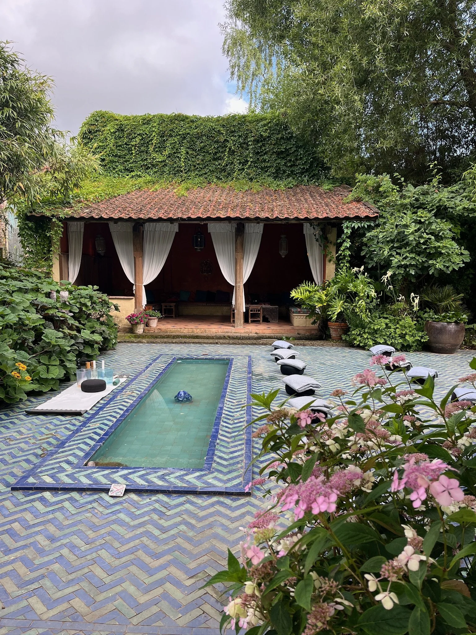 A backyard scene with a small rectangular swimming pool on a patterned tiled surface, surrounded by lush greenery and flowering plants. In the background, there's a covered outdoor lounge area with white curtains, chairs, and a wooden sofa. Pink flowers are in the foreground.