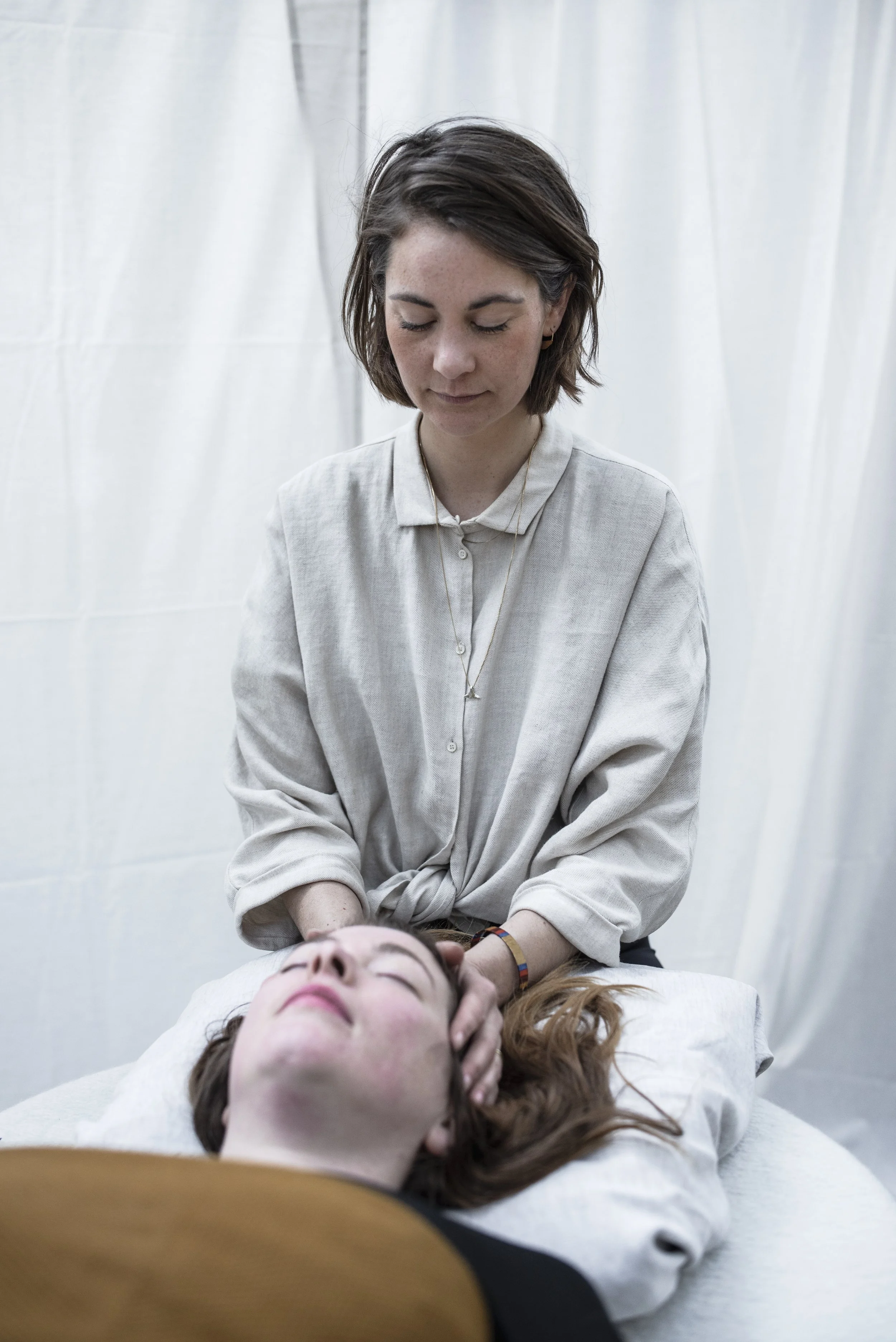 A woman giving a neck massage to another woman lying on a massage table, with a plain white curtain background in Nijmegen
