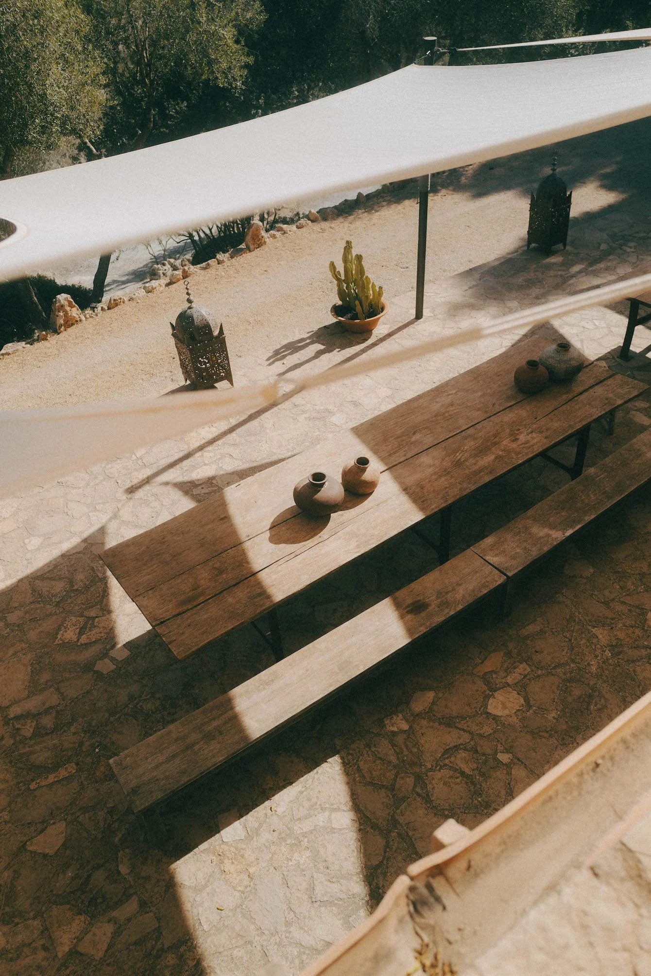 Twee houten banken op een terras met keramieken potten voor een retreat in Mallorca, een cactus in een pot, sierlijke lantaarns en een witte parasol, met een tuin met bomen op de achtergrond.