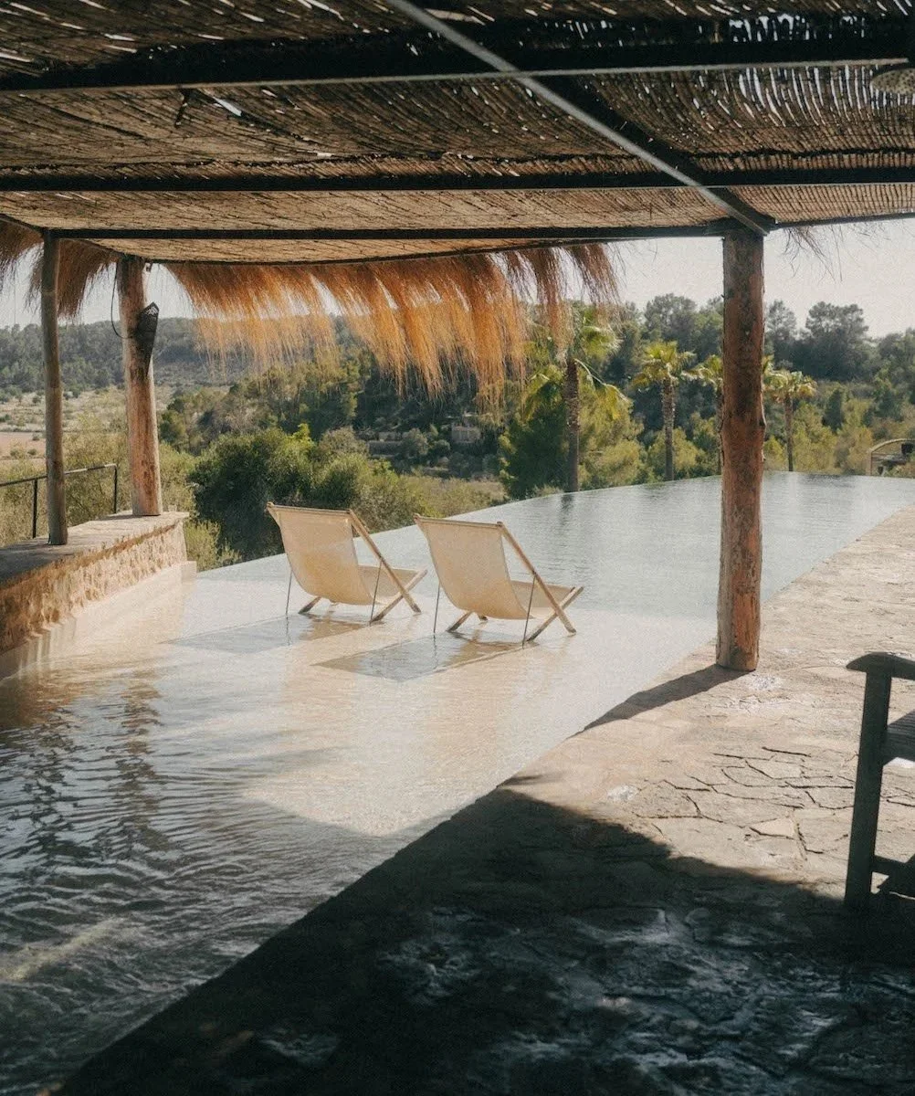 Twee ligbedden naast een infinity pool met uitzicht op een landschap met bomen en palmbomen van Mallorca, onder een bungalowdak met natuurlijke materialen.