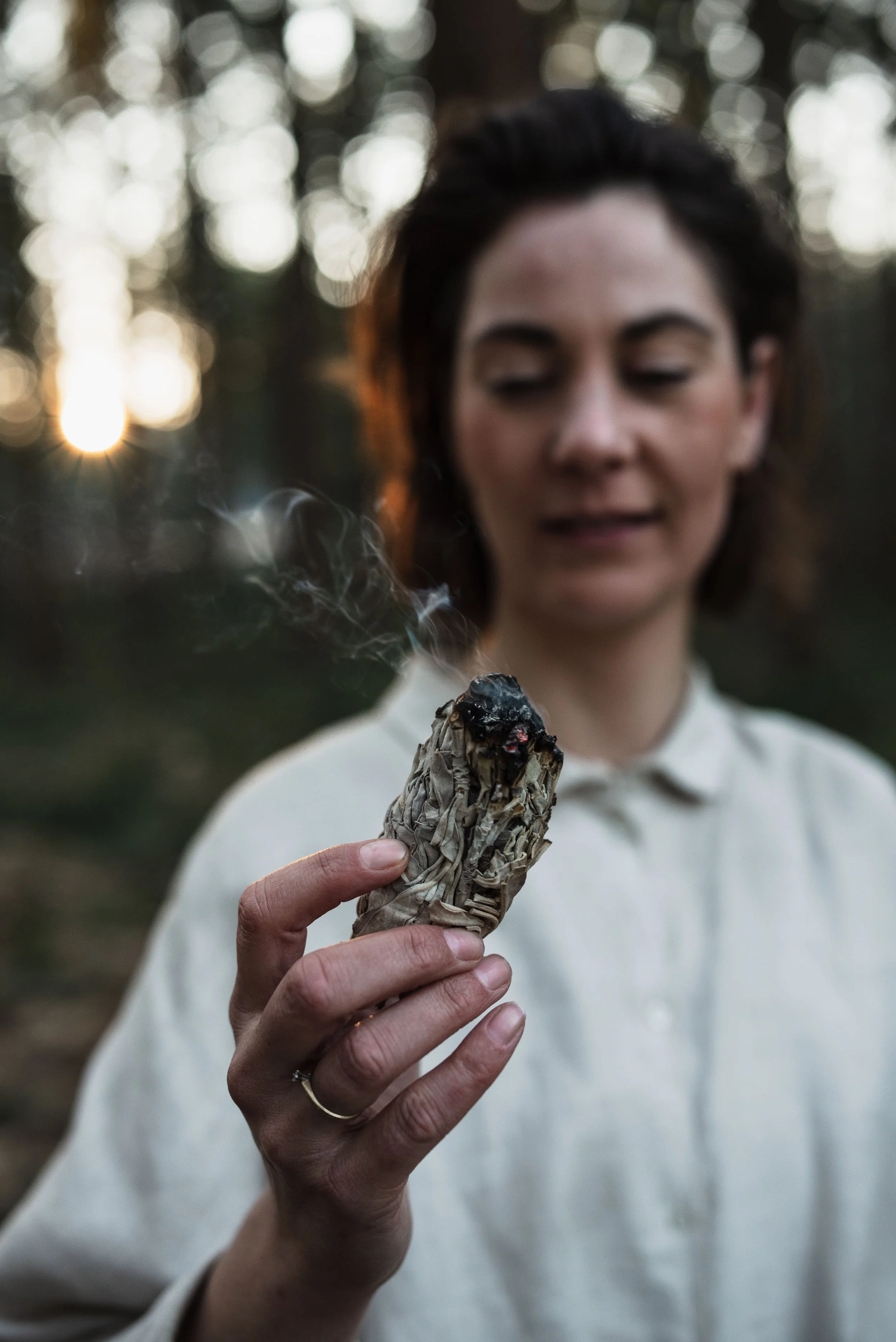 A woman holding a smoking smudge stick or bundle in her hand, with a blurred outdoor forest background at sunset at Nijmegen.