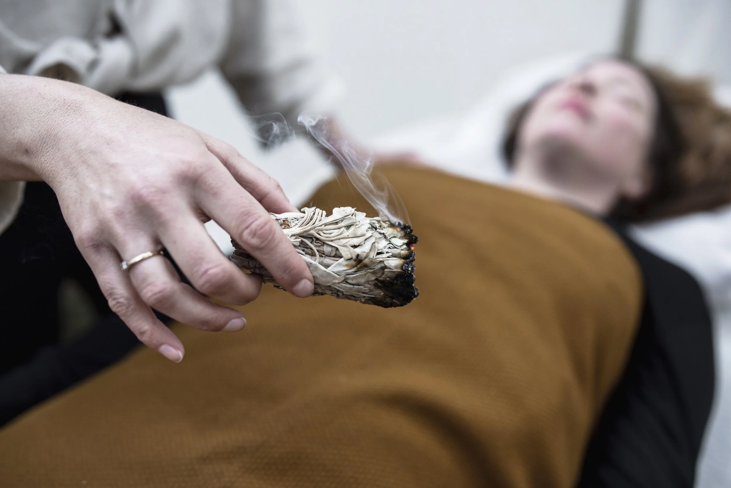 Person holding a burning stage of sage near a woman lying on a bed with eyes closed in Oosterhout