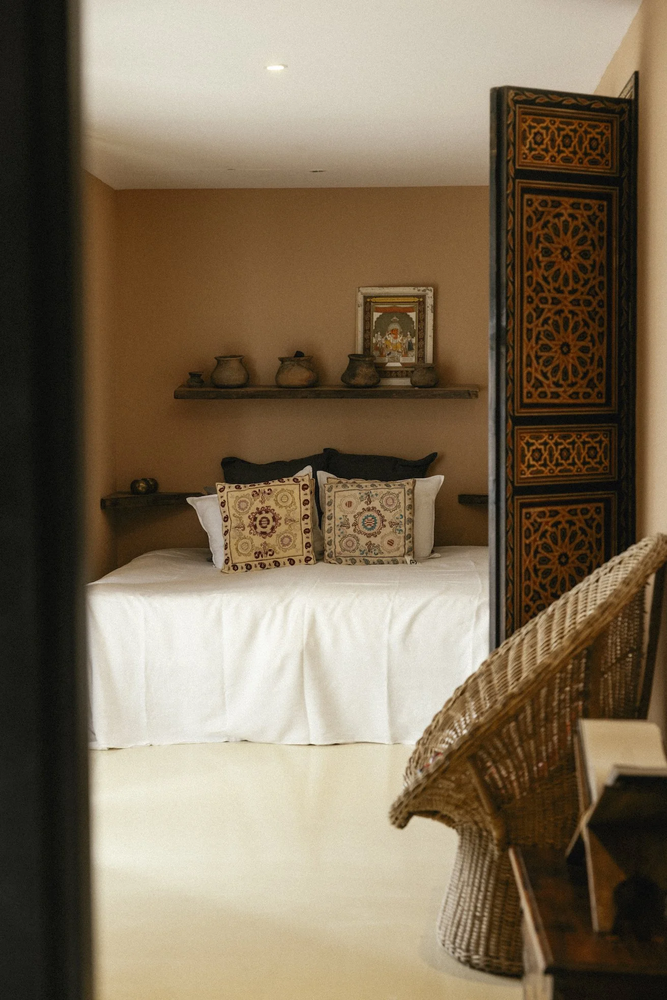 Een slaapkamer met een bed in Osa Major, Mallorca, versierd met vijf kussens met patronen, een houten muurplank met potten en een afbeelding, en een houten kamerscherm met oranje en zwarte patronen, een rieten stoel links.