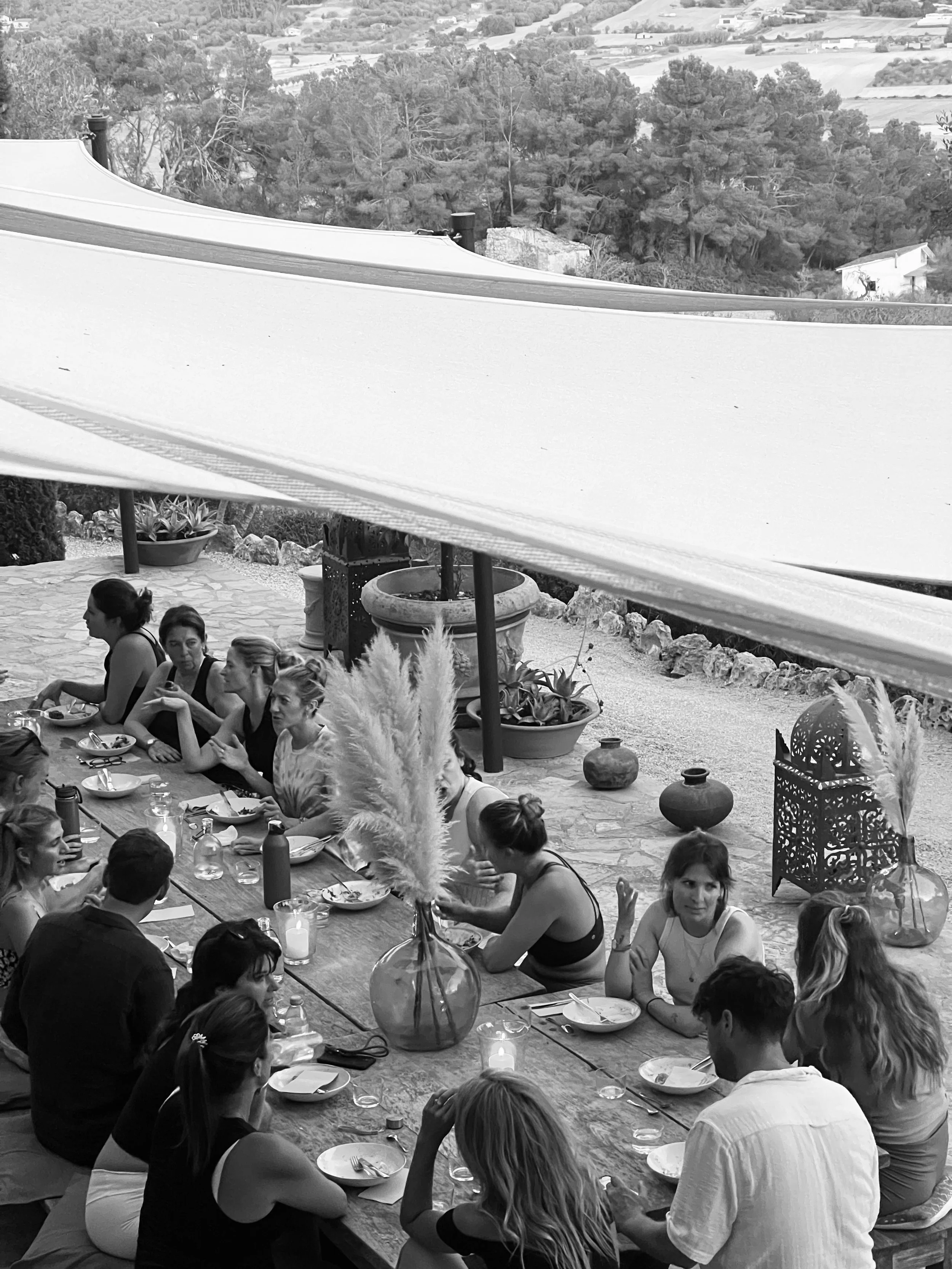 Een groep mensen zit aan een lange tafel buiten in Osa Major, Mallorca, lijken te genieten van een maaltijd, omgeven door planten en decoratieve lantaarns, met een landschap op de achtergrond.