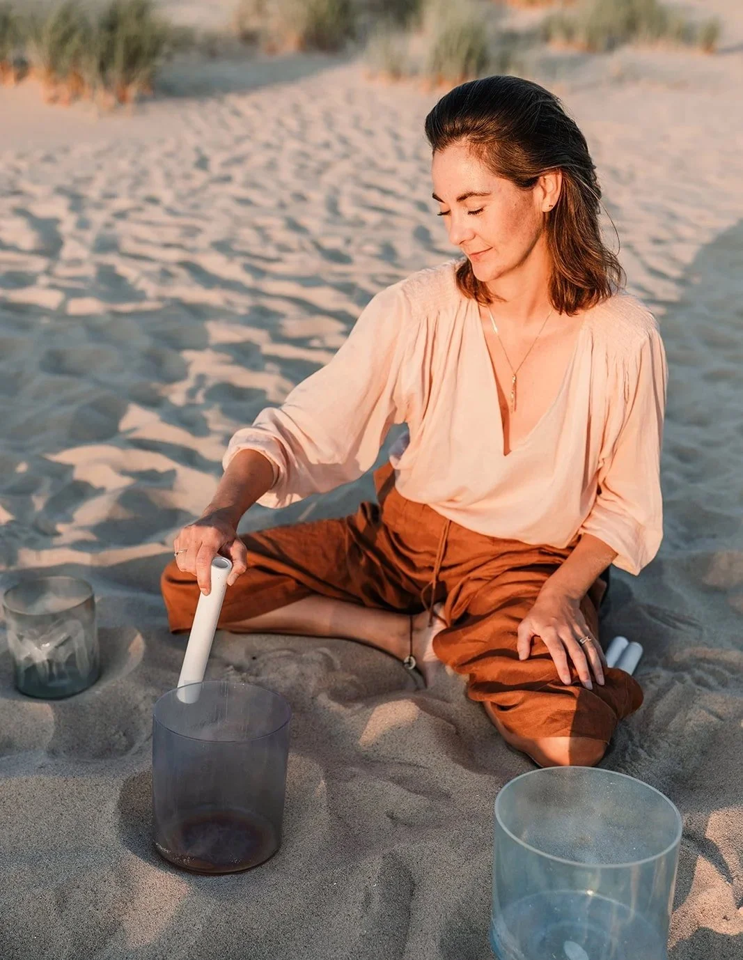 Vrouw zit op het zand op het strand en speelt met een glazen apparaat, mogelijk een muziekinstrument of een wetenschappelijk instrument, met andere glazen containers rondom haar.