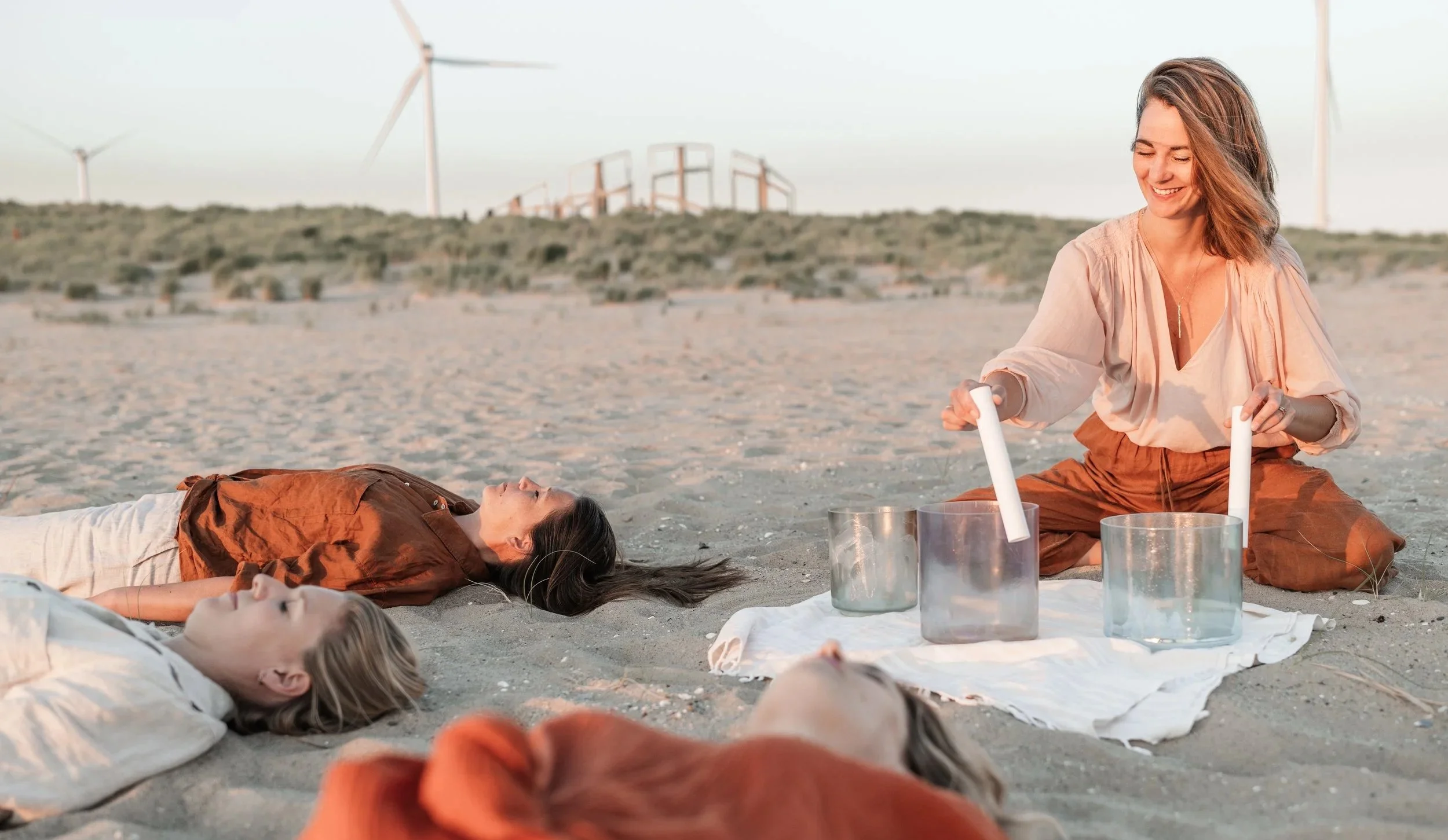Four women in a yoga or meditation session on the beach during sunset, with wind turbines in the background. Two women are lying down while one is playing crystal singing bowls.
