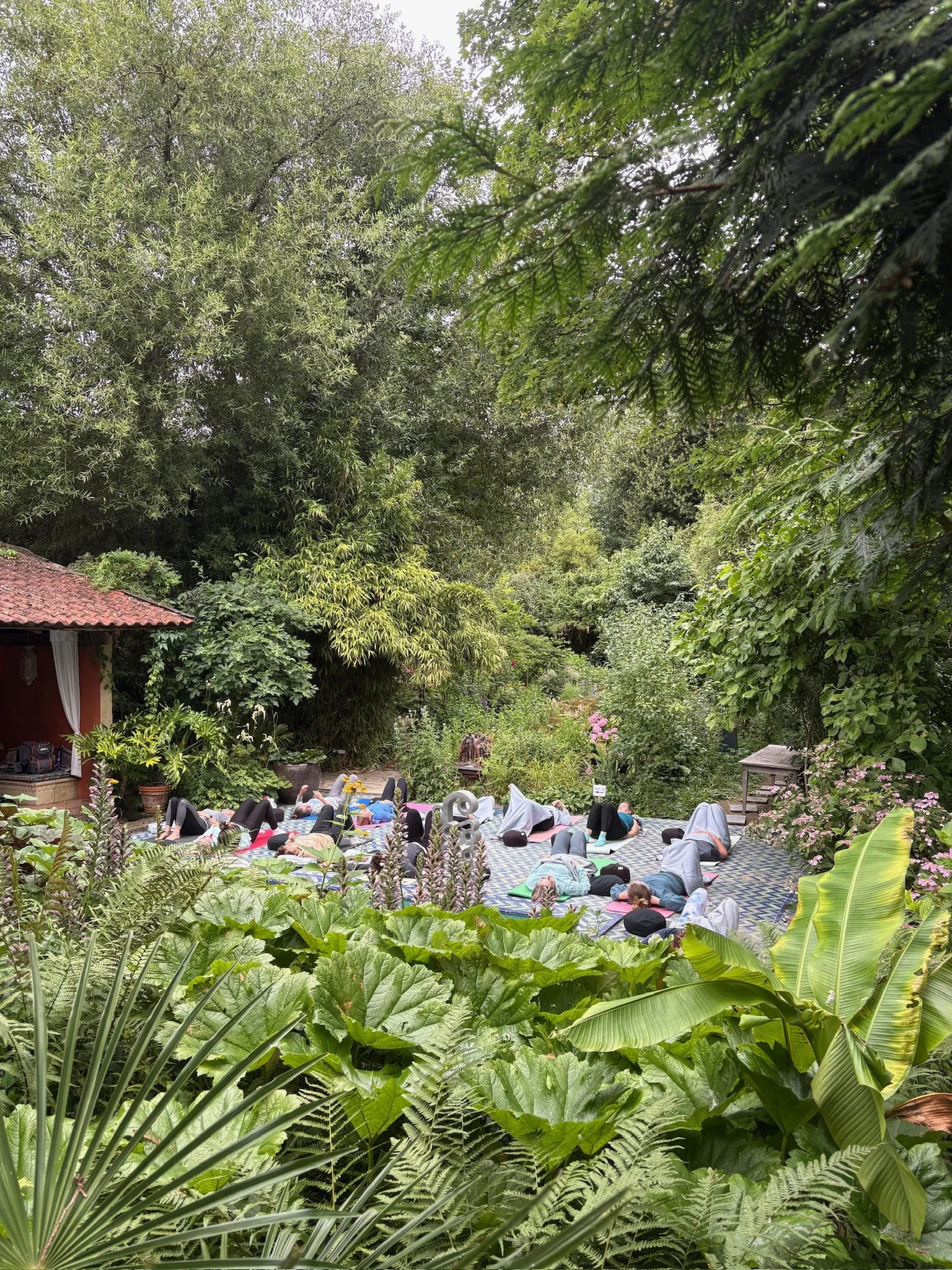 Mensen die yoga doen op matten in een tuin omringd door groene planten en bomen.