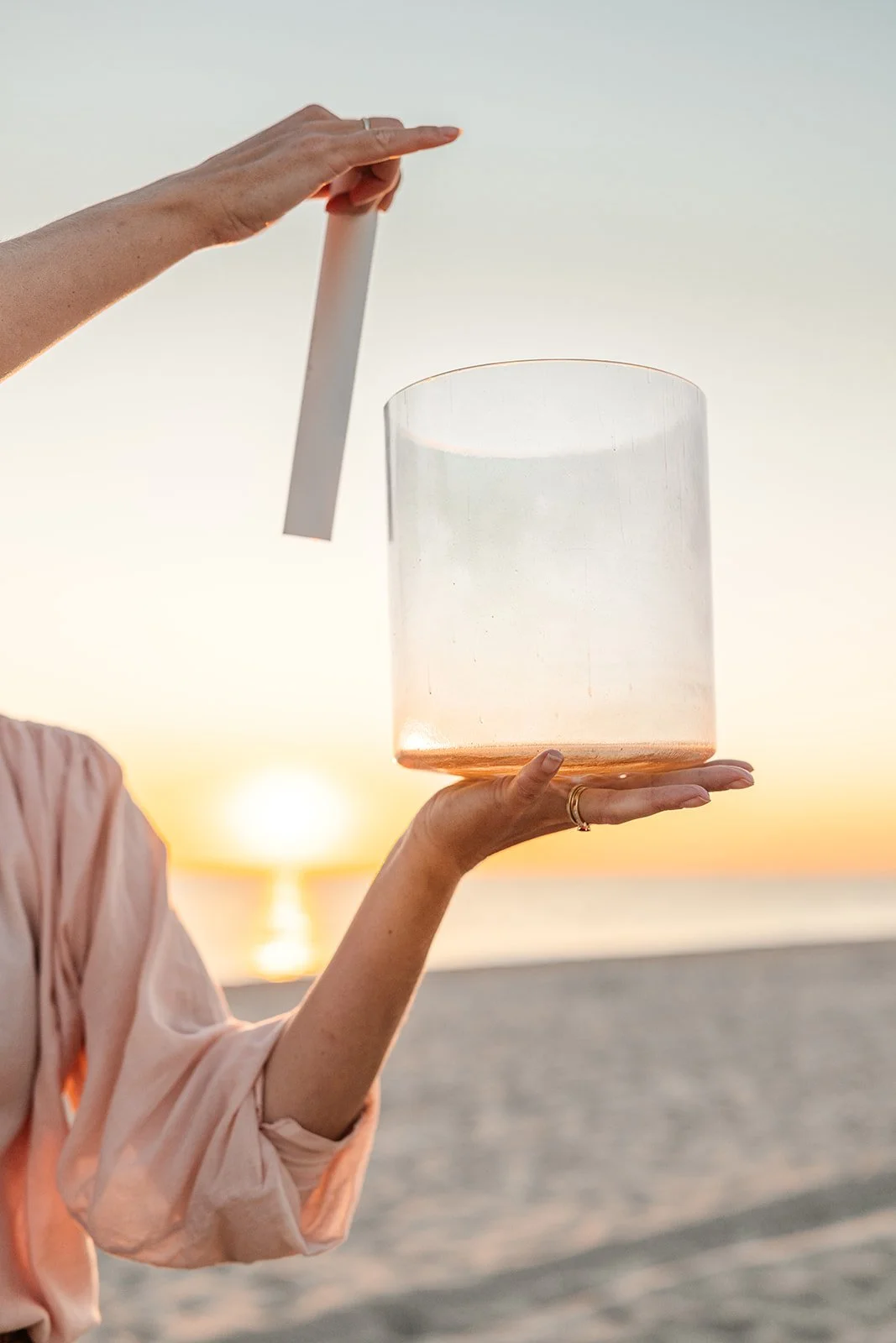 Person holding a crystal bowl with a white rectangular object inside, near the beach at sunset.