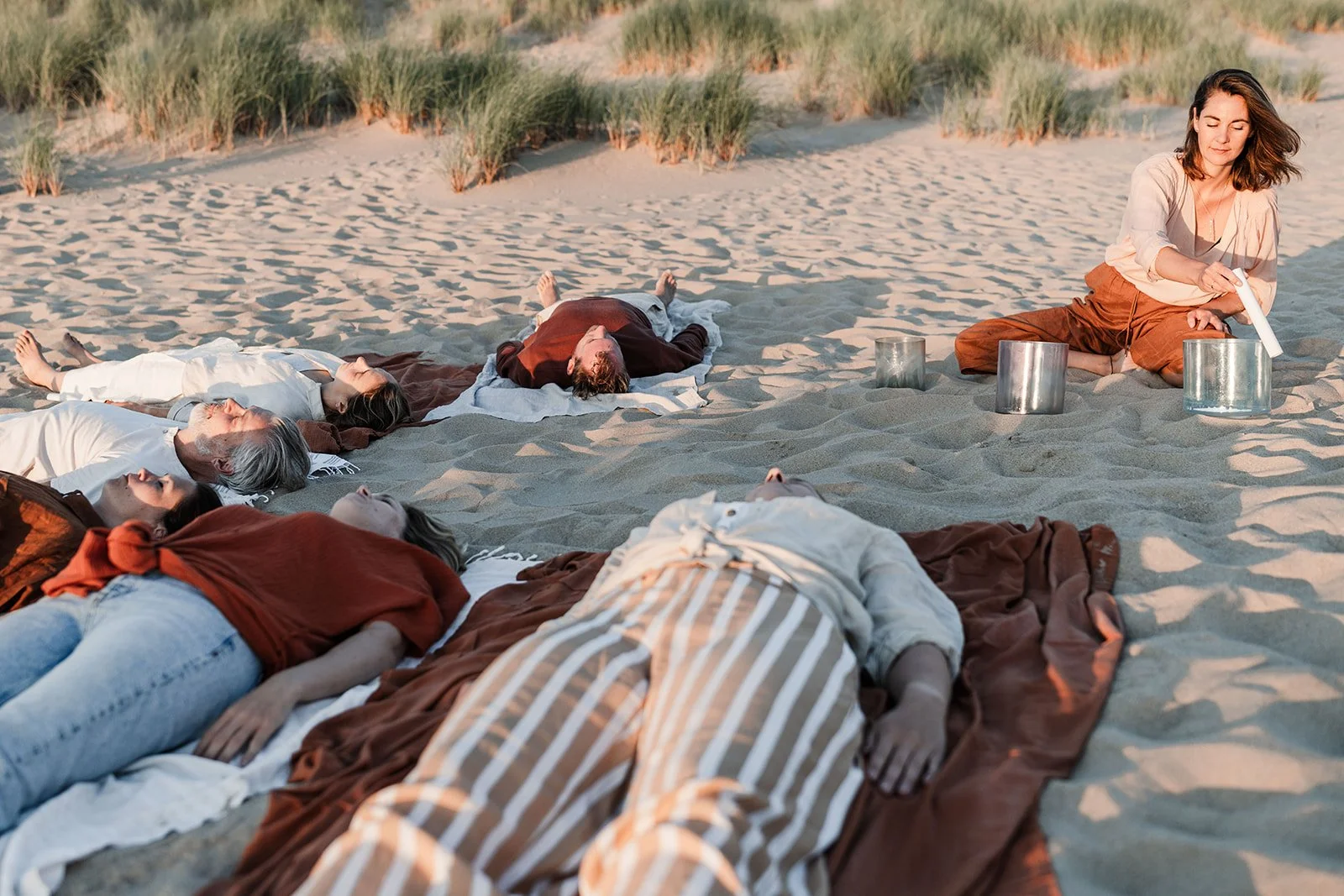 Group of people lying on the sandy beach with their eyes closed, while a woman is sitting and playing singing bowls.