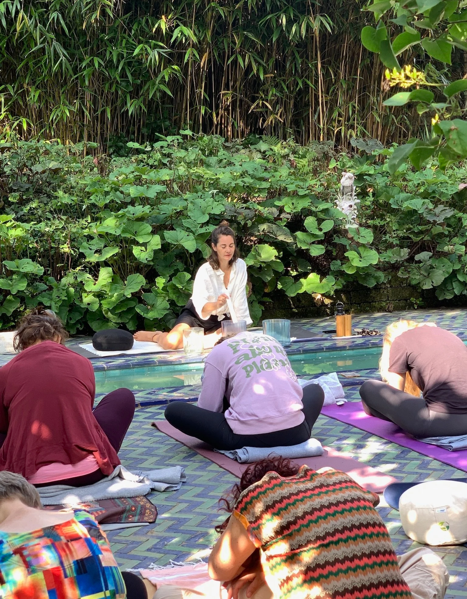 Mensen die deelnemen aan een yoga- of meditatieklas in een tuin met groene planten, lezend uit boeken en zittend op matten.