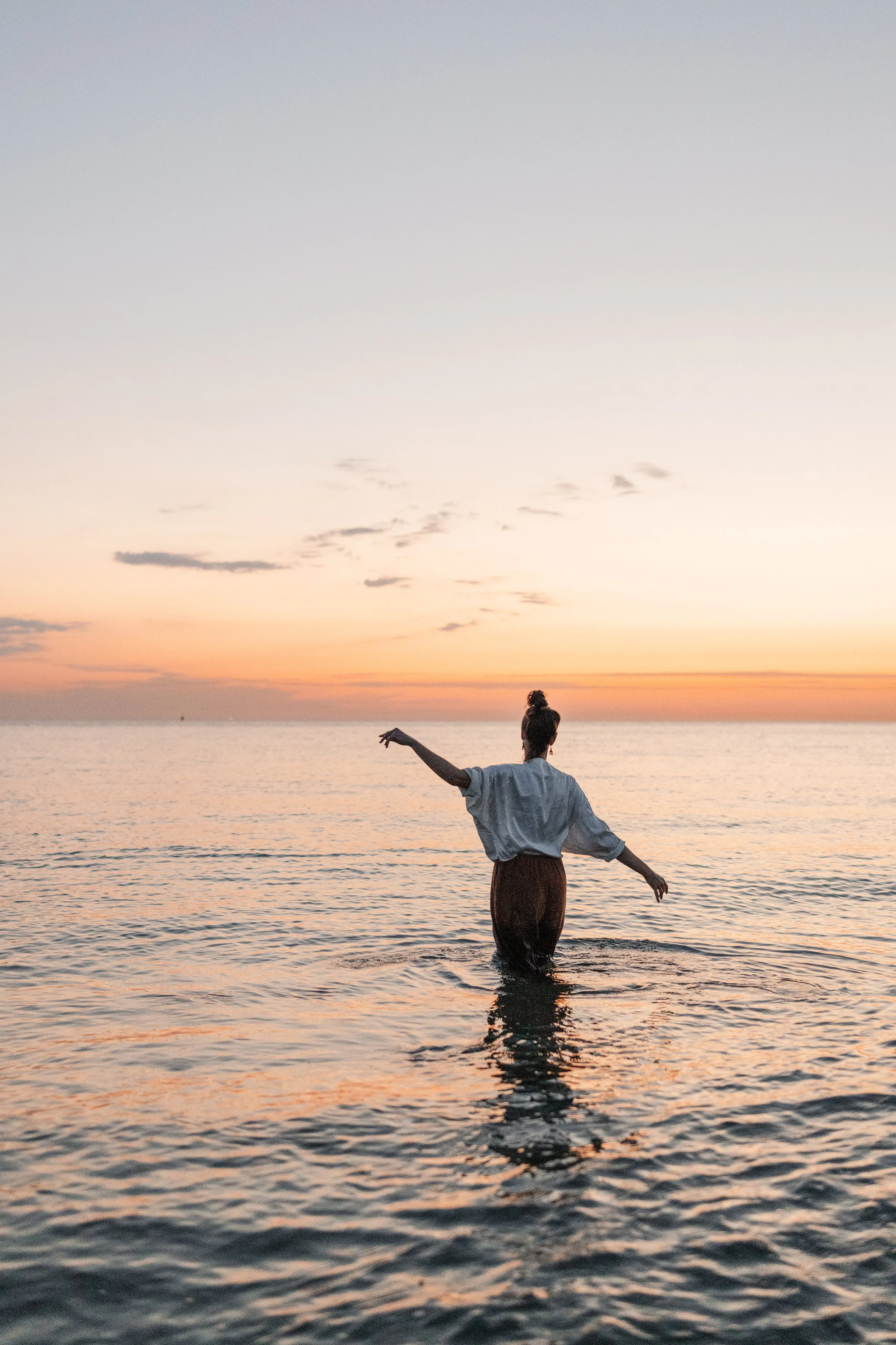 Een persoon staat in de zee tijdens zonsondergang met armen uitgestrekt en kijkt naar de horizon.