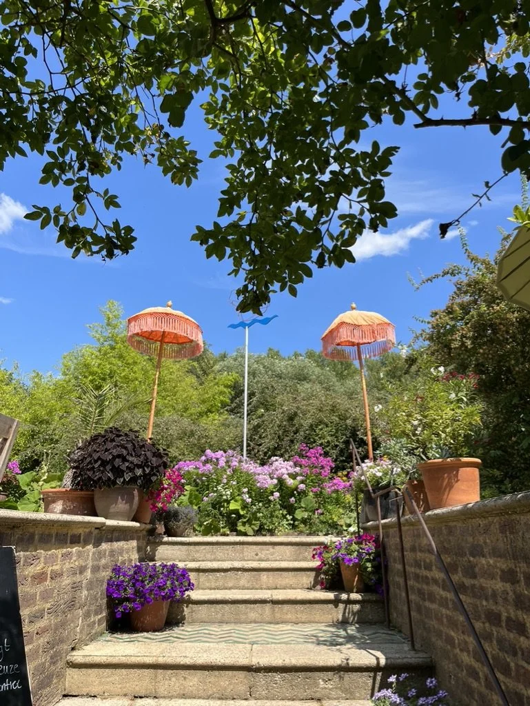 Stairs leading up to a garden with colorful flower pots and pink umbrellas, surrounded by lush greenery under a blue sky.
