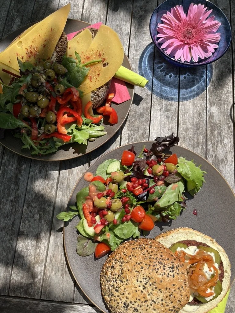Een afbeelding van twee borden met salade, een sandwich en een glas water met een roze bloem erin, op een houten tafel in de zon.