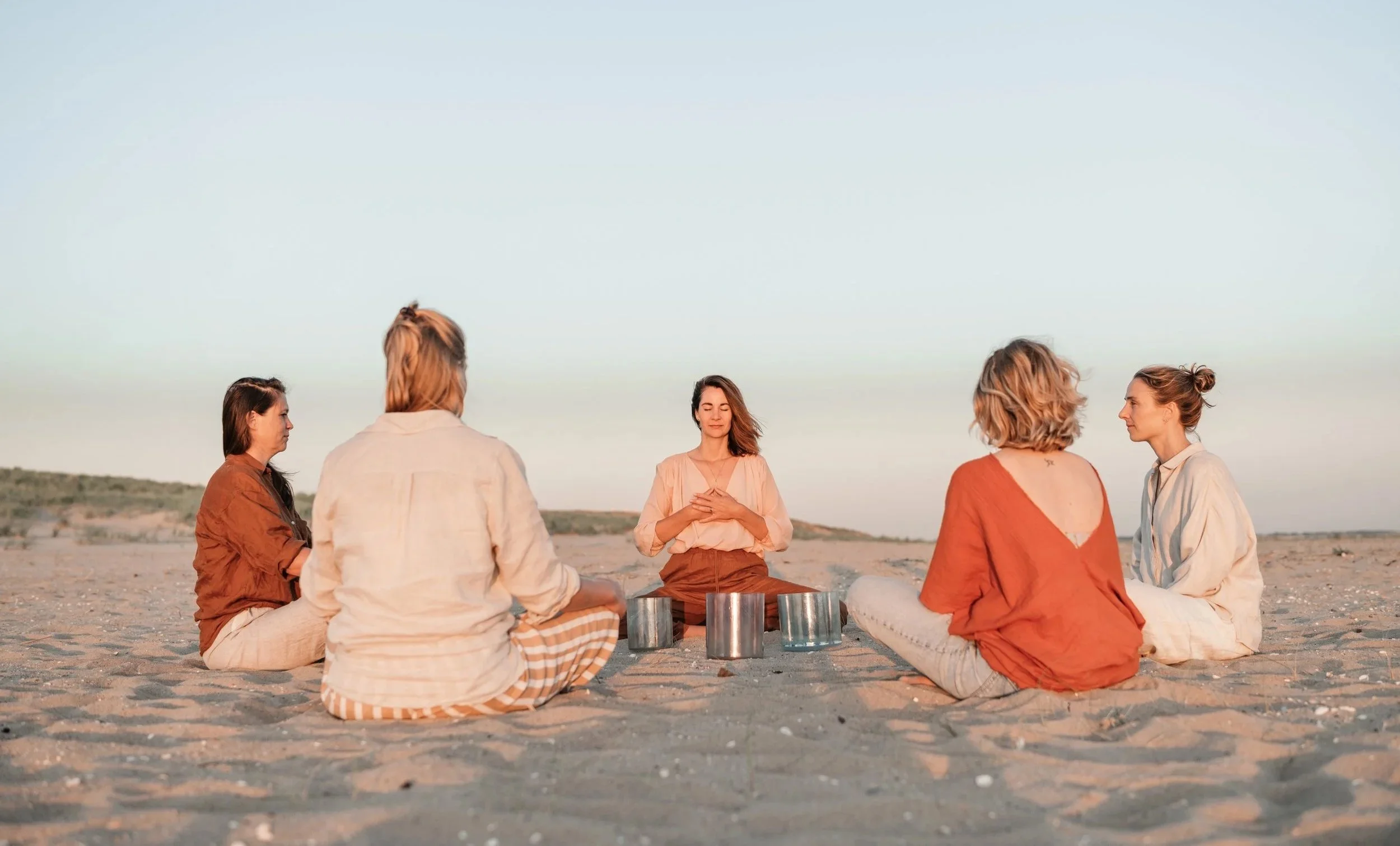 Six women sitting in a circle on a sandy beach with wind turbines in the background, enjoying a peaceful moment during sunset or sunrise.