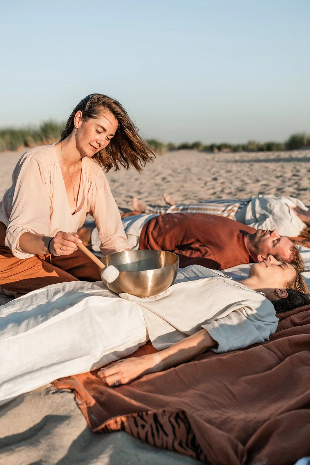 A woman plays a crystal singing bowl on a blanket at the beach while a man and woman relax nearby on the sand at sunset.