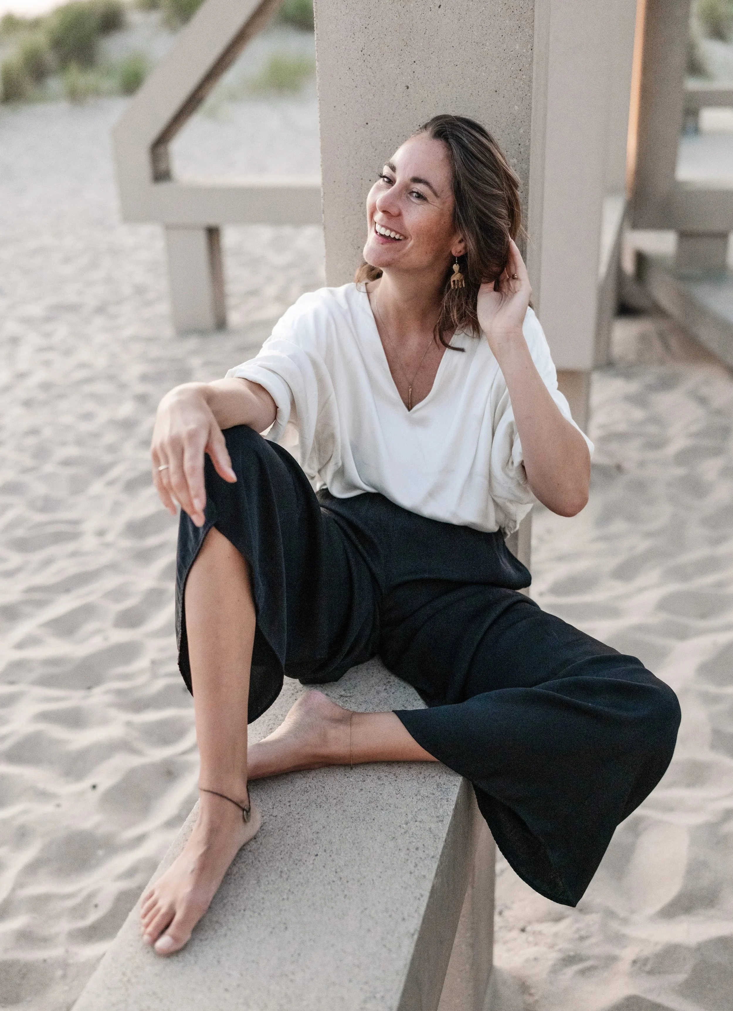 Een lachende vrouw die naast een betonnen balustrade op het strand zit, sportieve kleding en oorbellen.