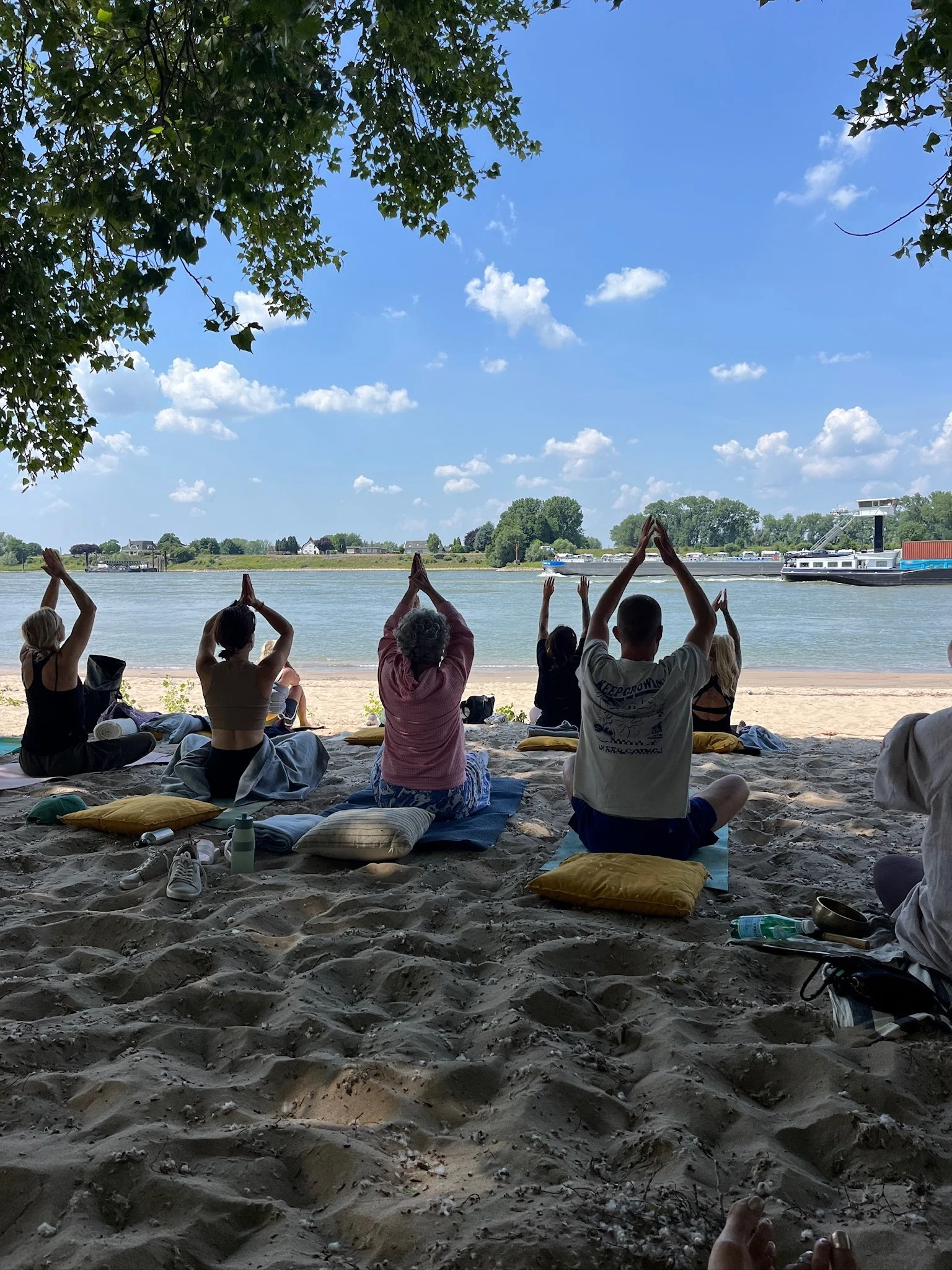 People practicing yoga on a sandy beach near a river under a tree on a sunny day at Millinger Thee Tuin.