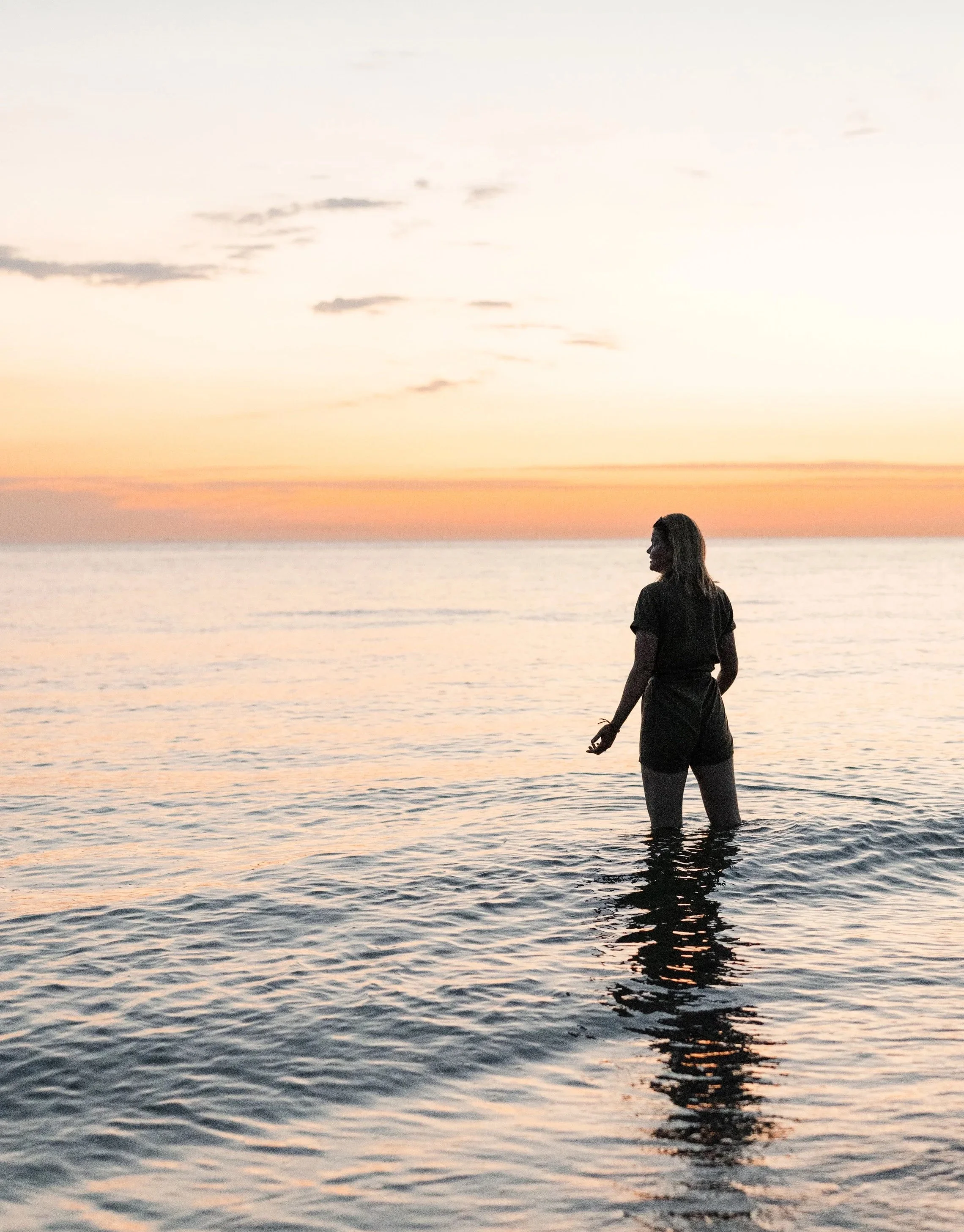 Een vrouw staat in de zee, met de horizon en een kleurrijke zonsondergang op de achtergrond.