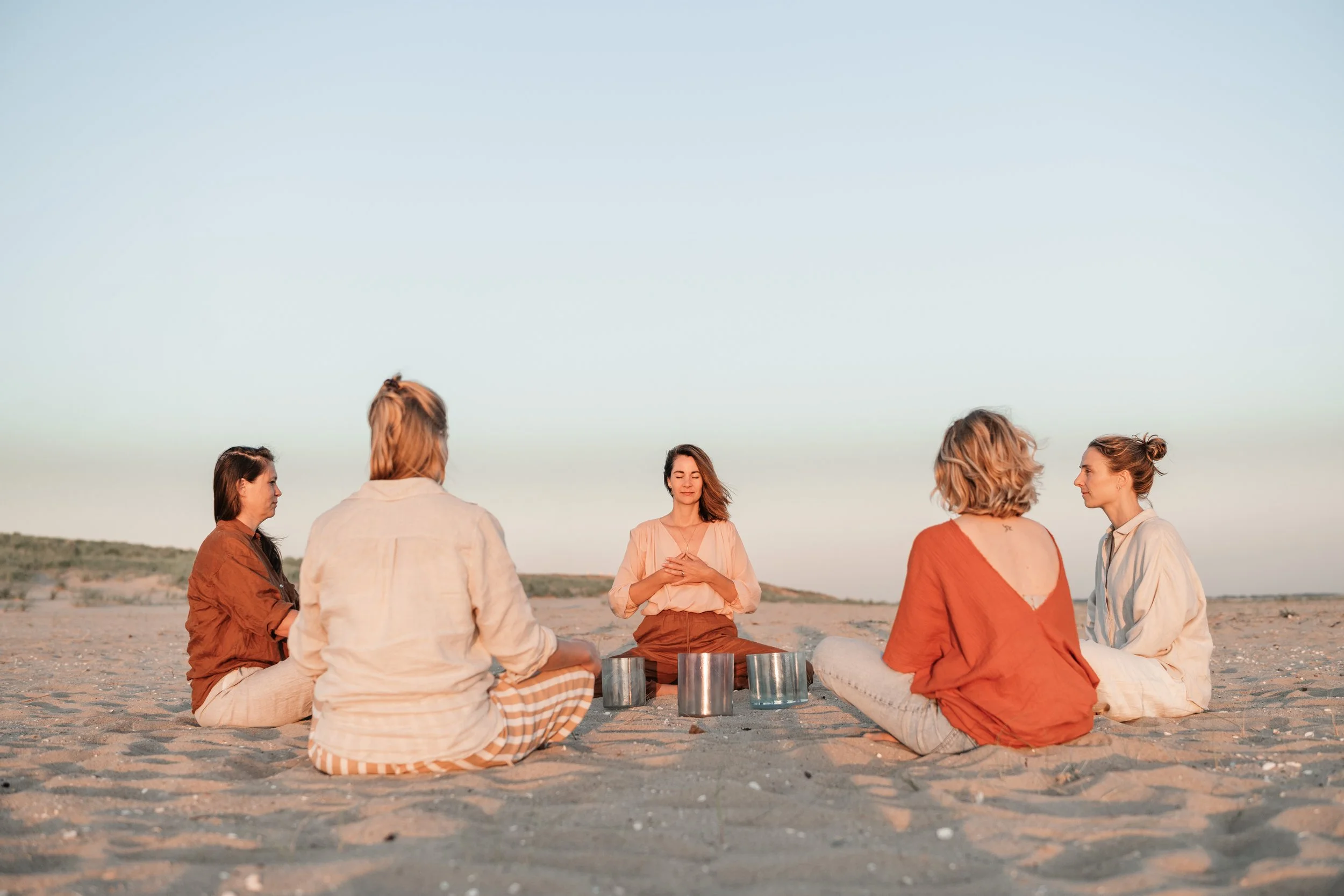 Six women sitting in a circle on the sand at the beach during sunset, with one woman in the middle playing crystal singing bowls.