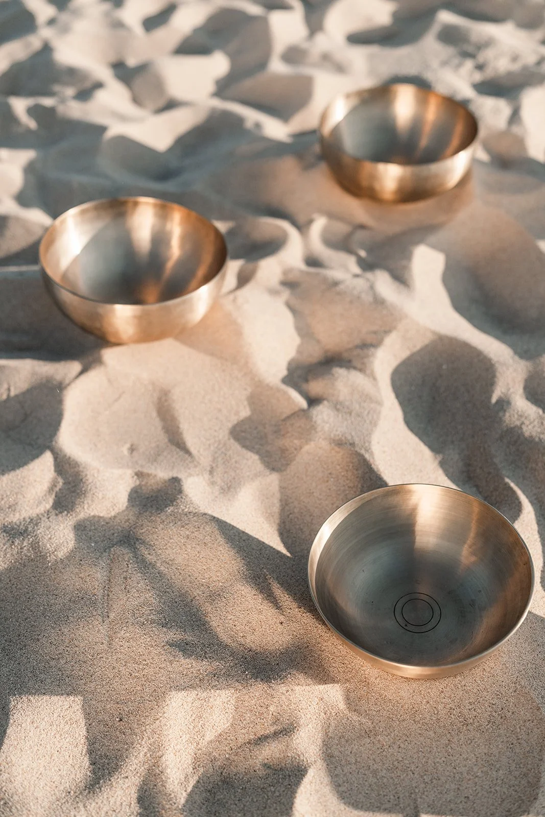 Twee metalen schalen en een kom liggen op het zandstrand, met verzengende zonlicht en schaduw.