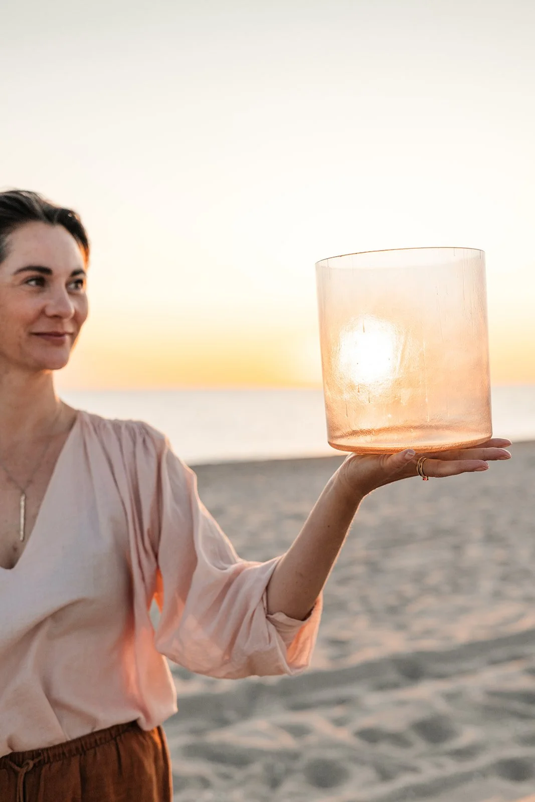 A woman holding a large glass lamp on the beach during sunset.