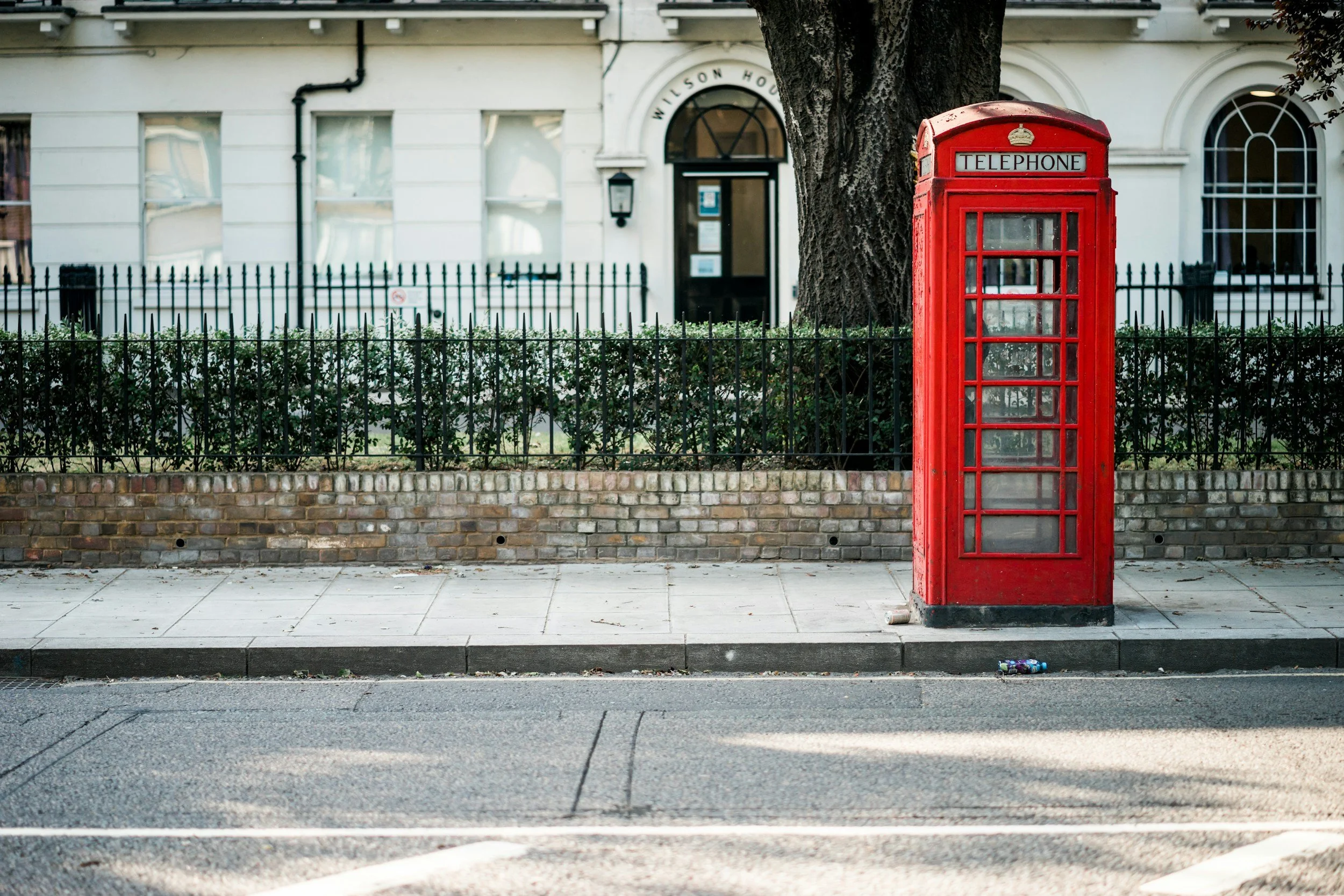 Red British telephone booth on sidewalk next to a tree and a black iron fence in front of a white building with arched windows.