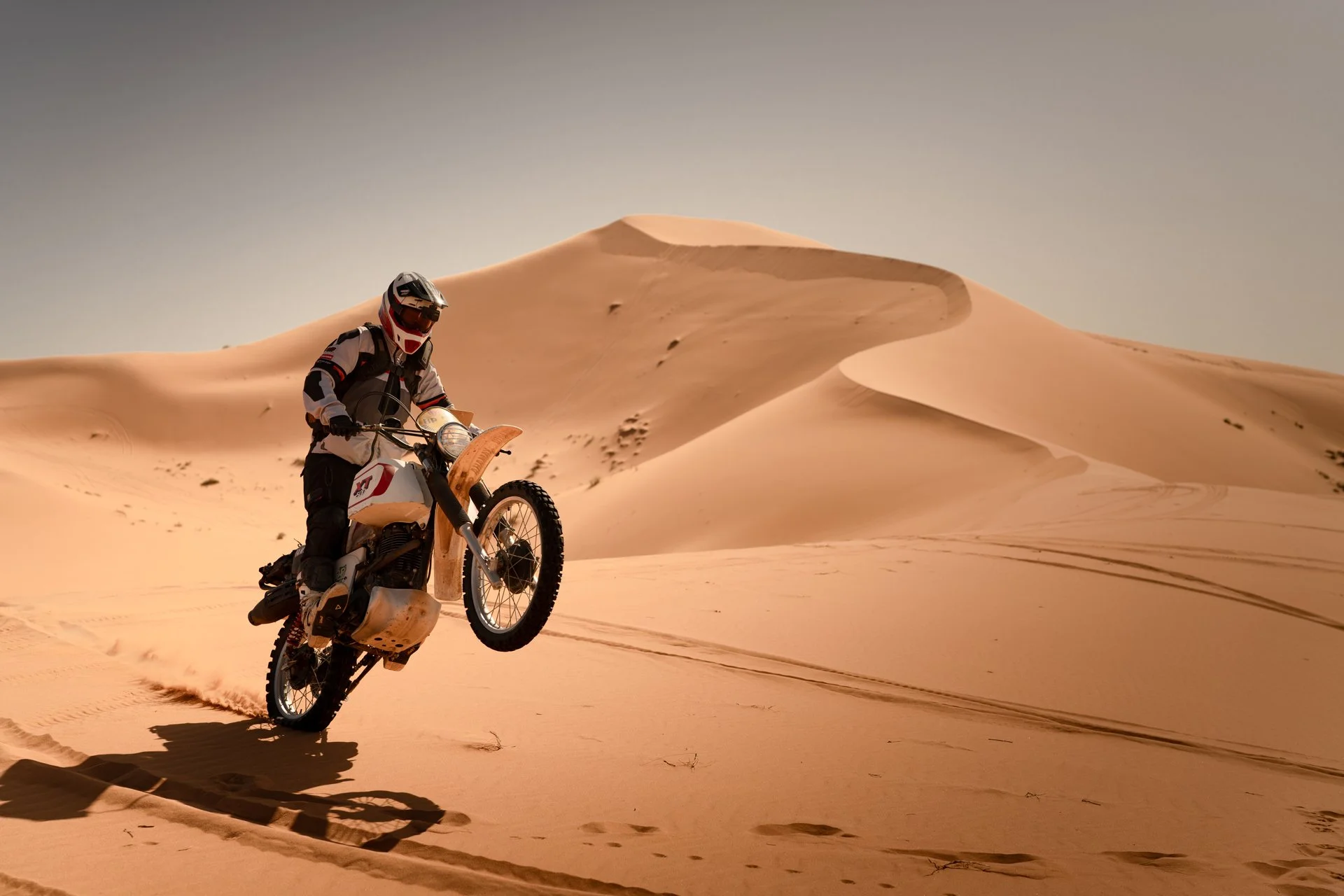 Motorcyclist performing a wheelie on a vintage dirt bike XT 500 in the desert with sand dunes in the background.  Shot from the Brake Magazine film 100 Kicks in the Desert. Shot from the Brake Magazine film 100 Kicks in the Desert. © Richard Kurowski