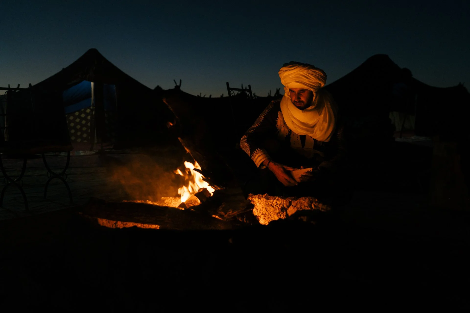 A man dressed in traditional Indian attire, including a turban and scarf, sitting near a campfire at night with tents in the background.  Shot from the Brake Magazine film 100 Kicks in the Desert. Shot from the Brake Magazine film 100 Kicks in the De