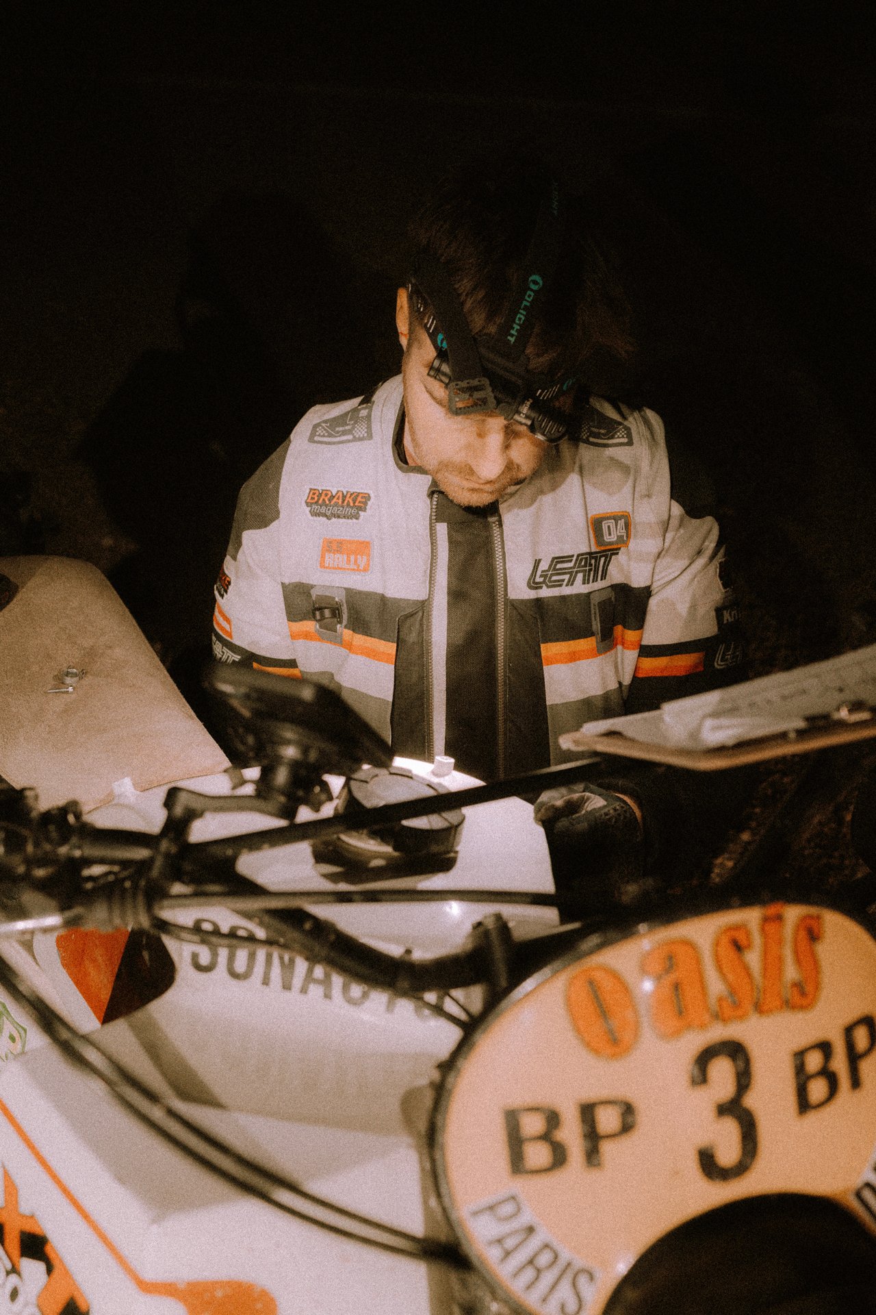 Llewellyn Pavey wearing a Leatt Rally 5.5  jacket with patches, a headlamp,  working a vintage Dakar Rally motorcycle at night. Shot from the Brake Magazine film 100 Kicks in the Desert. © Richard Kurowski