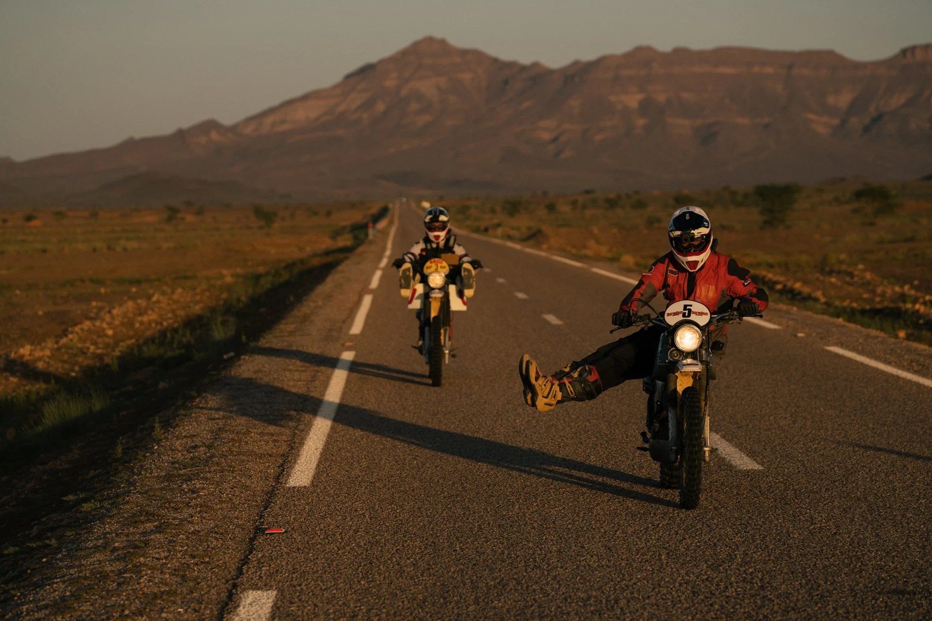 Two motorcyclists riding on a long empty road in a desert landscape with mountains in the background. Shot from the Brake Magazine film 100 Kicks in the Desert. © Richard Kurowski
