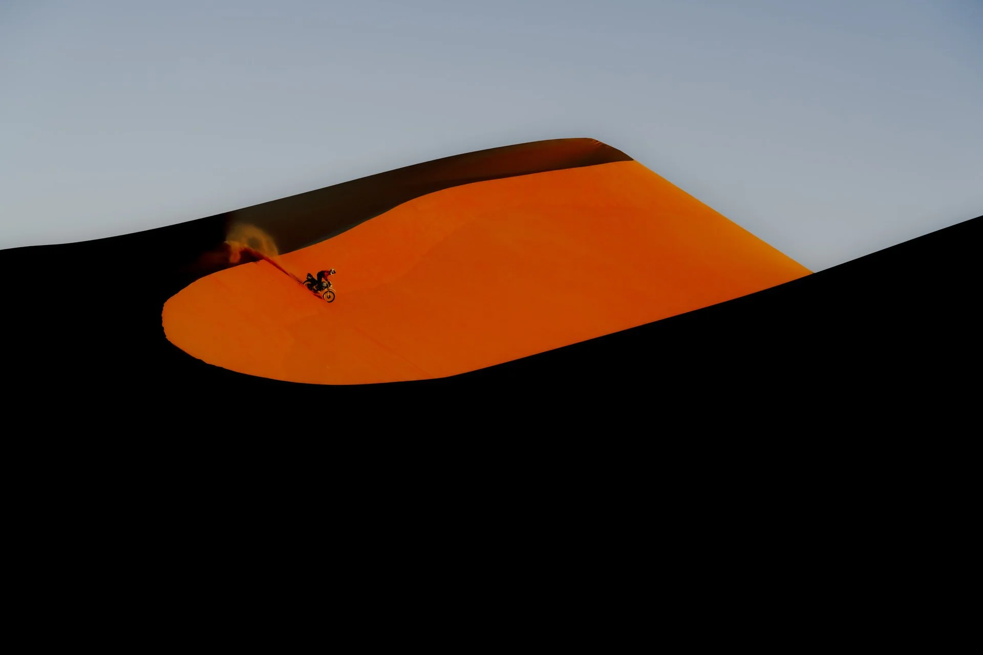 A person riding a motorcycle on a large sand dune in a desert, with a trail of dust behind them during sunrise.  Shot from the Brake Magazine film 100 Kicks in the Desert. Shot from the Brake Magazine film 100 Kicks in the Desert. © Richard Kurowski