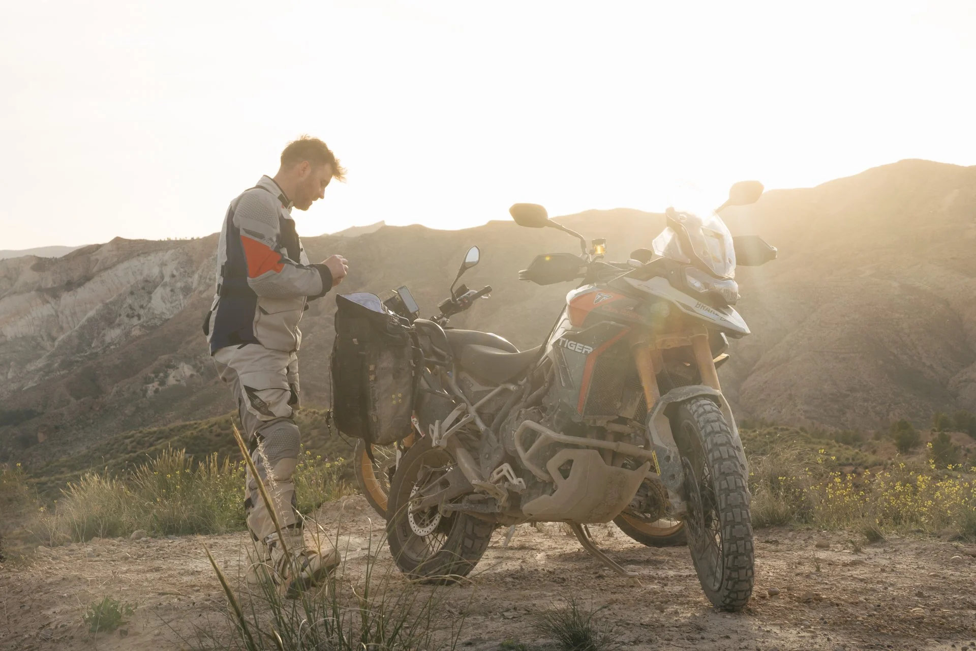 A man in adventure motorcycle gear standing next to an off-road motorcycle in a desert landscape during sunset. The man is Llel Pavey and the motorcycle is a Triumph Tiger 900. © Brake Magazine
