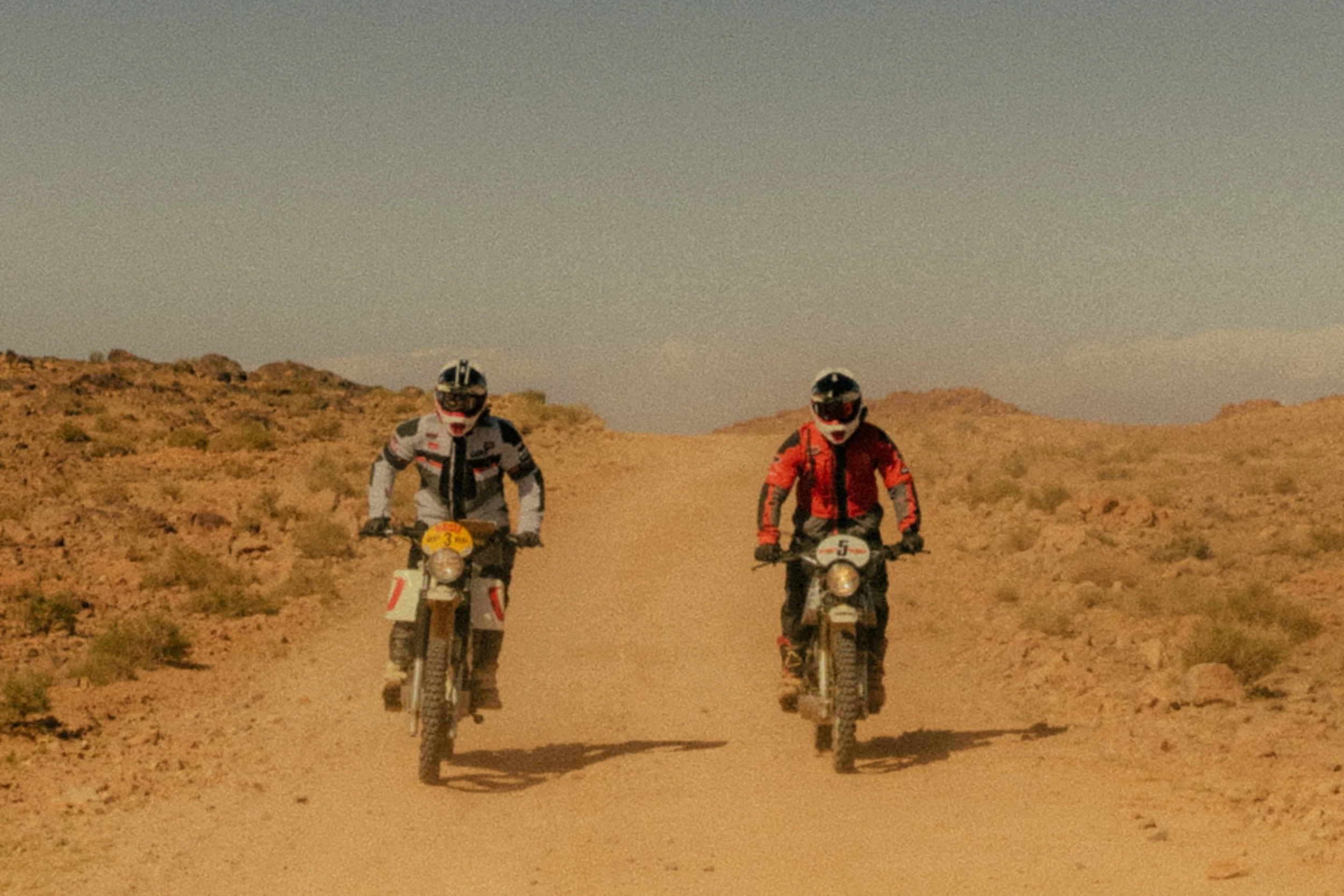 Two people riding vintage dirt bikes on a desert trail with rocky, sparse terrain and a clear sky. Shot from the Brake Magazine film 100 Kicks in the Desert. © Richard Kurowski