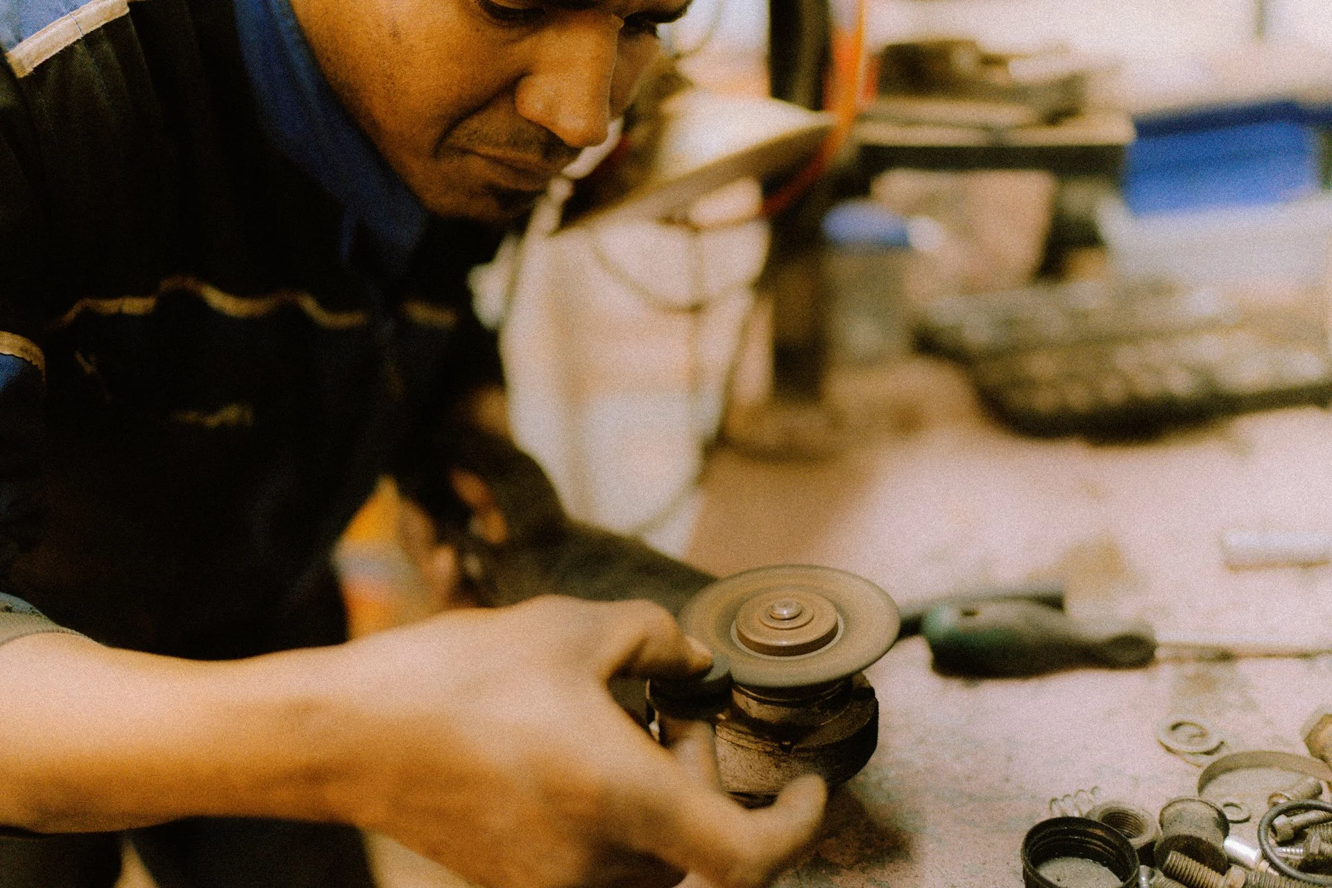 A man working with a grinding tool on a small mechanical part in a workshop, surrounded by various hardware components. Shot from the Brake Magazine film 100 Kicks in the Desert. © Richard Kurowski
