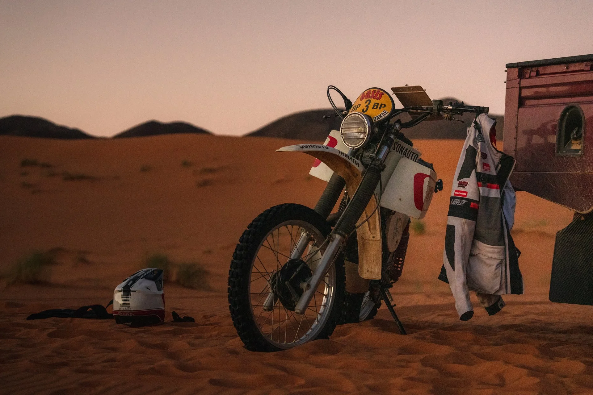 A vintage XT 500 Dakar Replica dirt bike with a headlight, parked on desert sand at dusk, with a helmet nearby and a jacket hanging on the bike, and dunes in the background.  Shot from the Brake Magazine film 100 Kicks in the Desert. © Richard Kurows