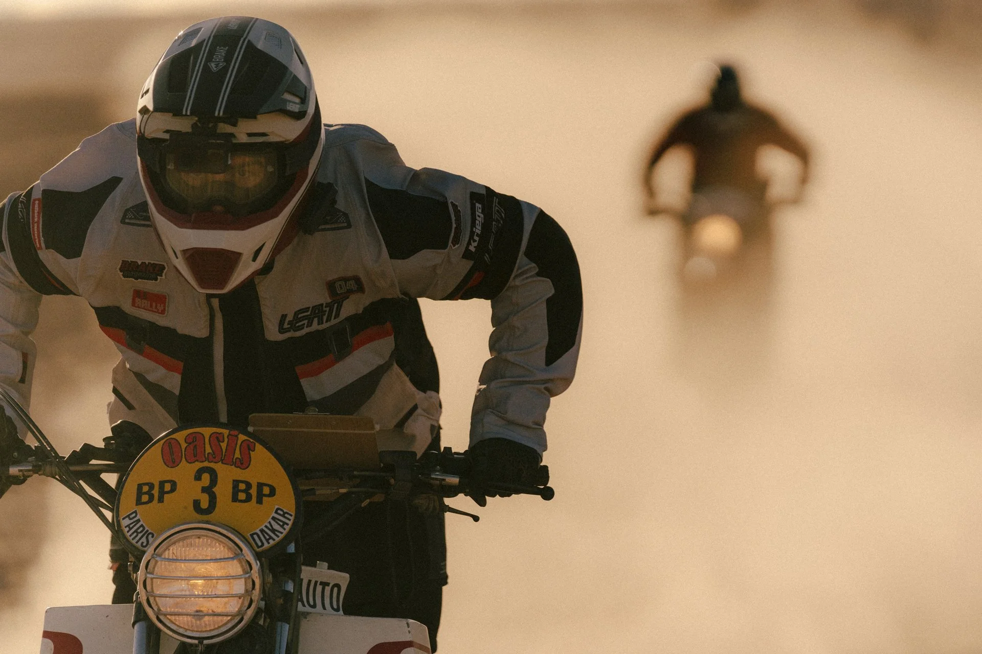 A person in a Leatt 5.5 Rally Jacket and 7.5 helmet riding a motorcycle through a desert, with another motorcycle rider blurred in the background. Shot from the Brake Magazine film 100 Kicks in the Desert. © Richard Kurowski