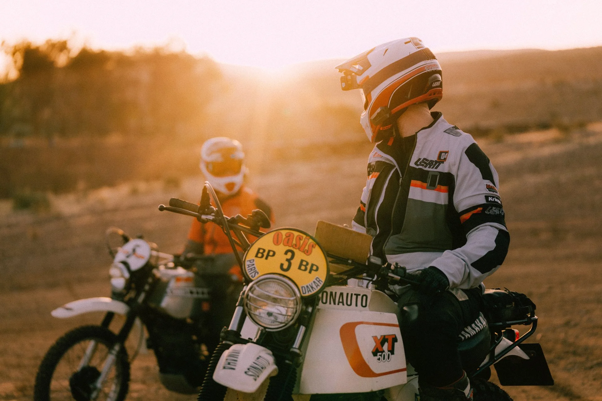 Two motorcyclists are riding dirt bikes in a desert landscape during sunset, wearing full gear and helmets. The gear is Leatt Rally 5.5.  Shot from the Brake Magazine film 100 Kicks in the Desert. © Richard Kurowski