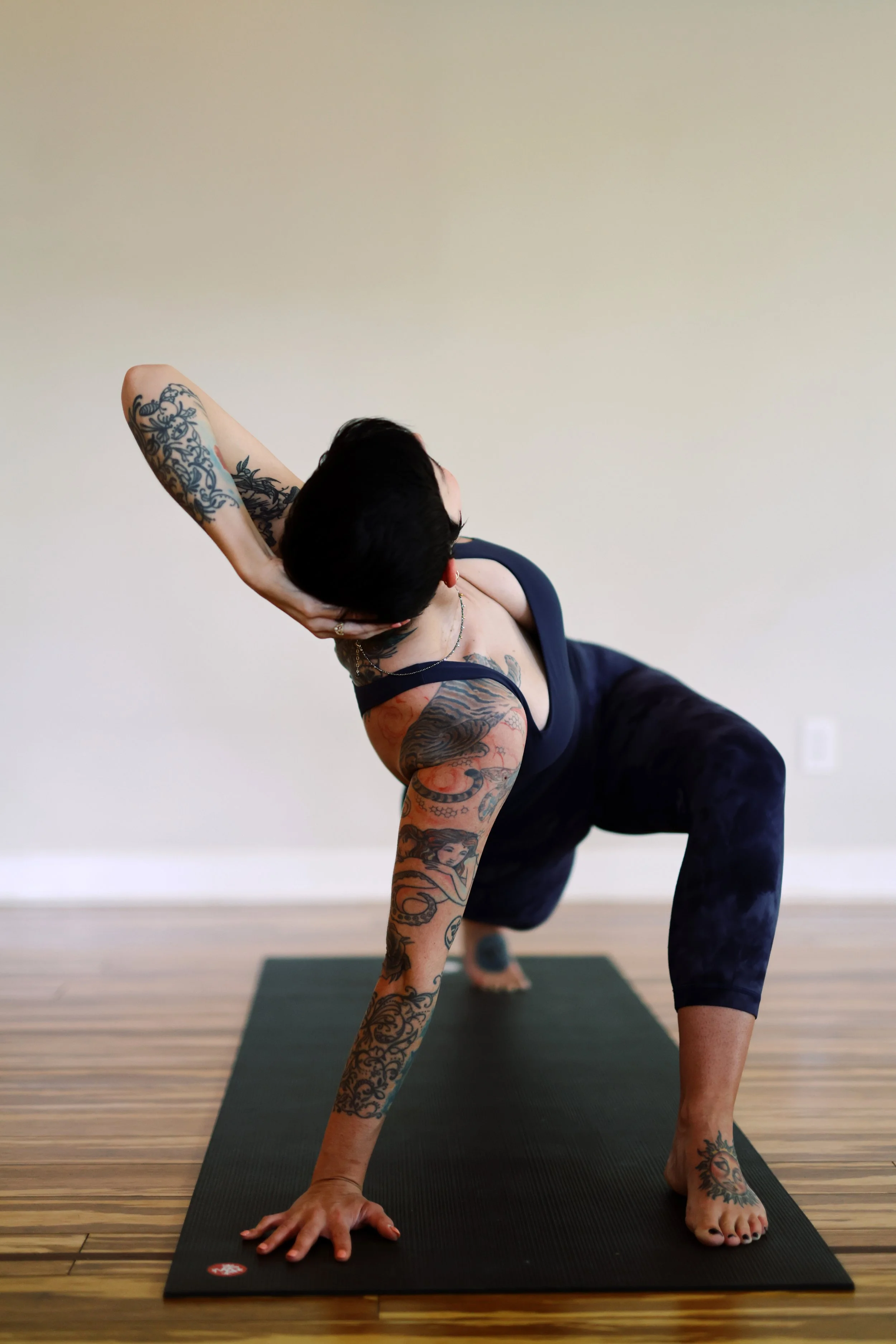 A person with tattoos doing a yoga pose on a black yoga mat with a wooden floor and blank wall background.